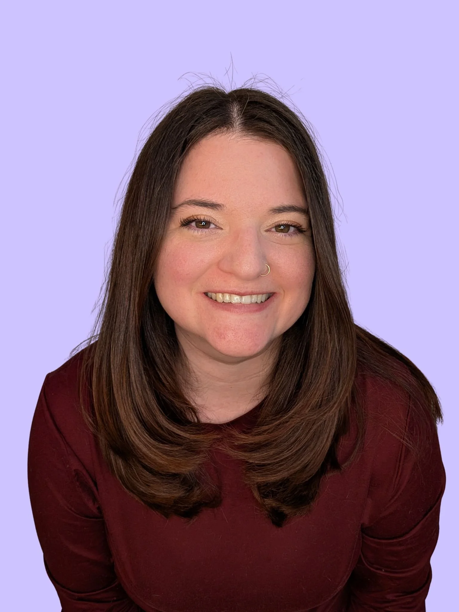 A woman with long brown hair, smiling, wearing a maroon top, posing against a light purple background.
