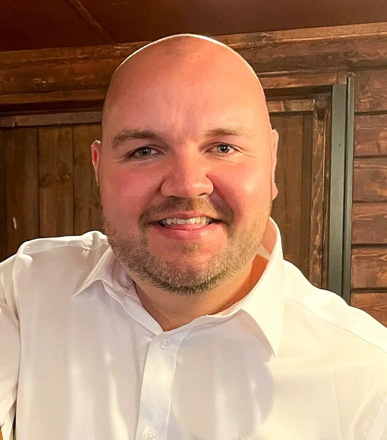 A smiling man with a shaved head and beard, wearing a white dress shirt, posing indoors with wooden walls in the background.
