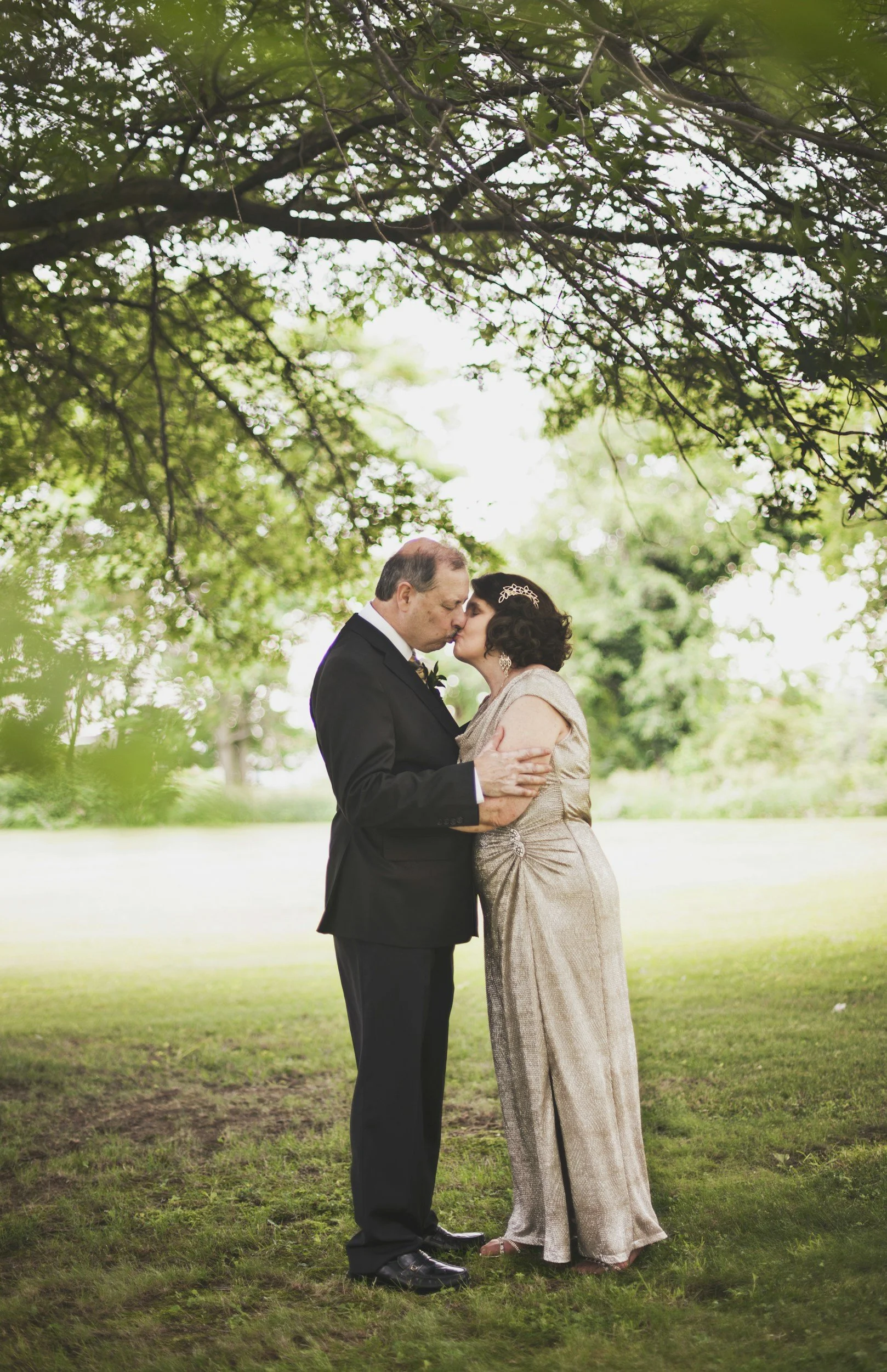 A middle-aged man and woman in wedding attire sharing a kiss outdoors under a canopy of tree branches and leaves.