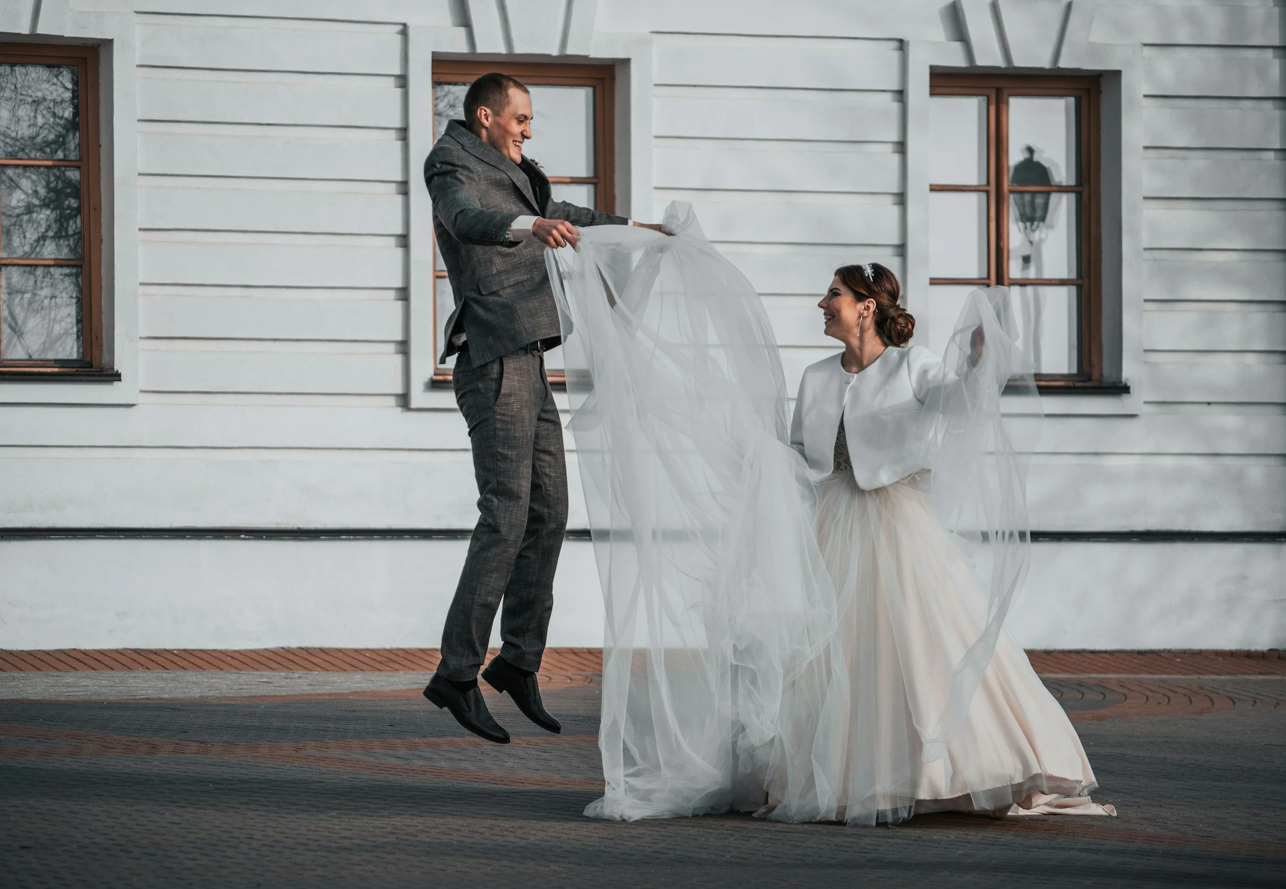 A bride with long red hair dancing with her eyes closed, holding a sheer veil, and a groom in a light gray jacket covering his face with a bouquet of white baby's breath flowers, standing against a dark wooden wall.