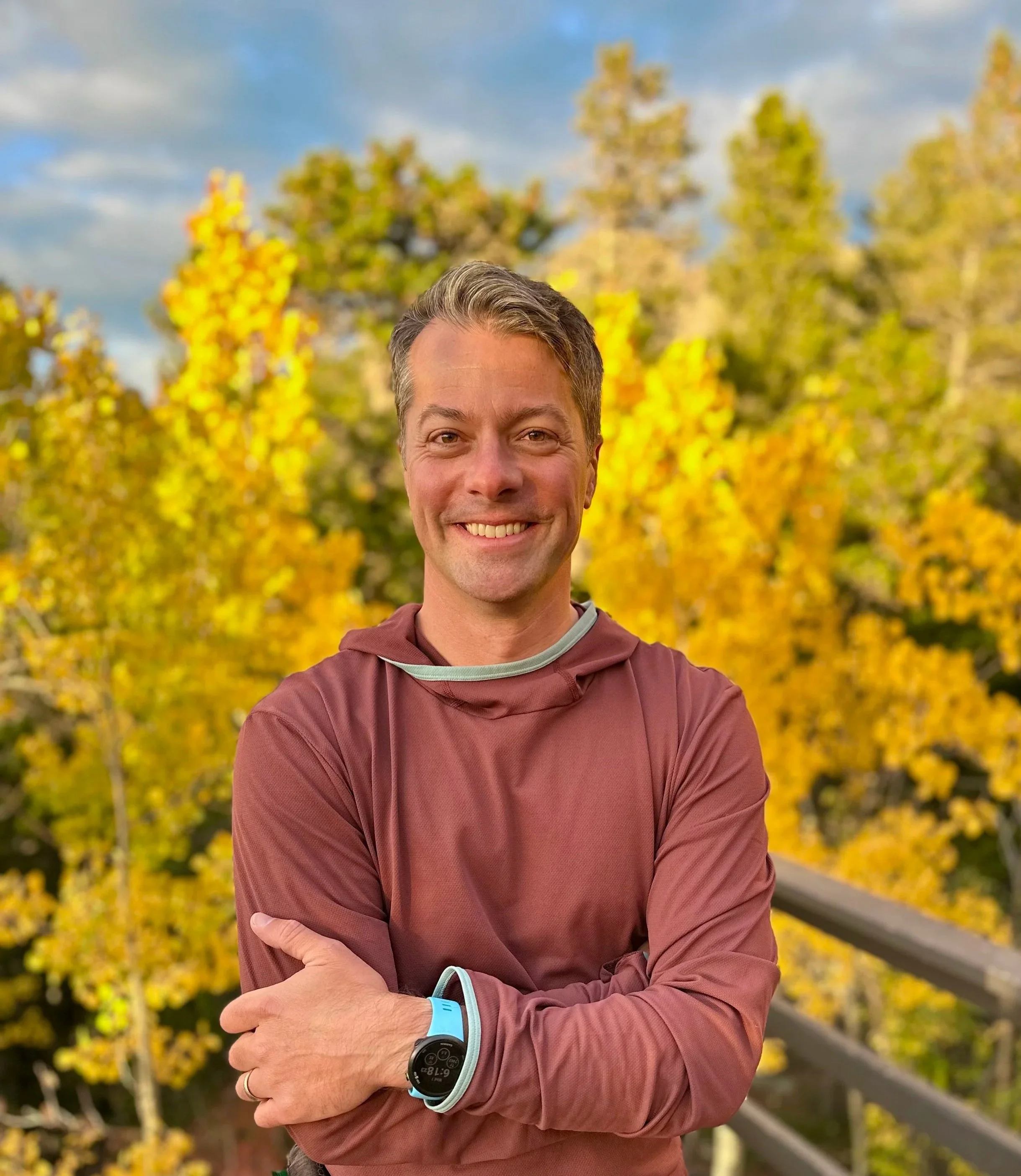A smiling man in a maroon athletic jacket standing outdoors on a wooden balcony with autumn yellow and green trees in the background.