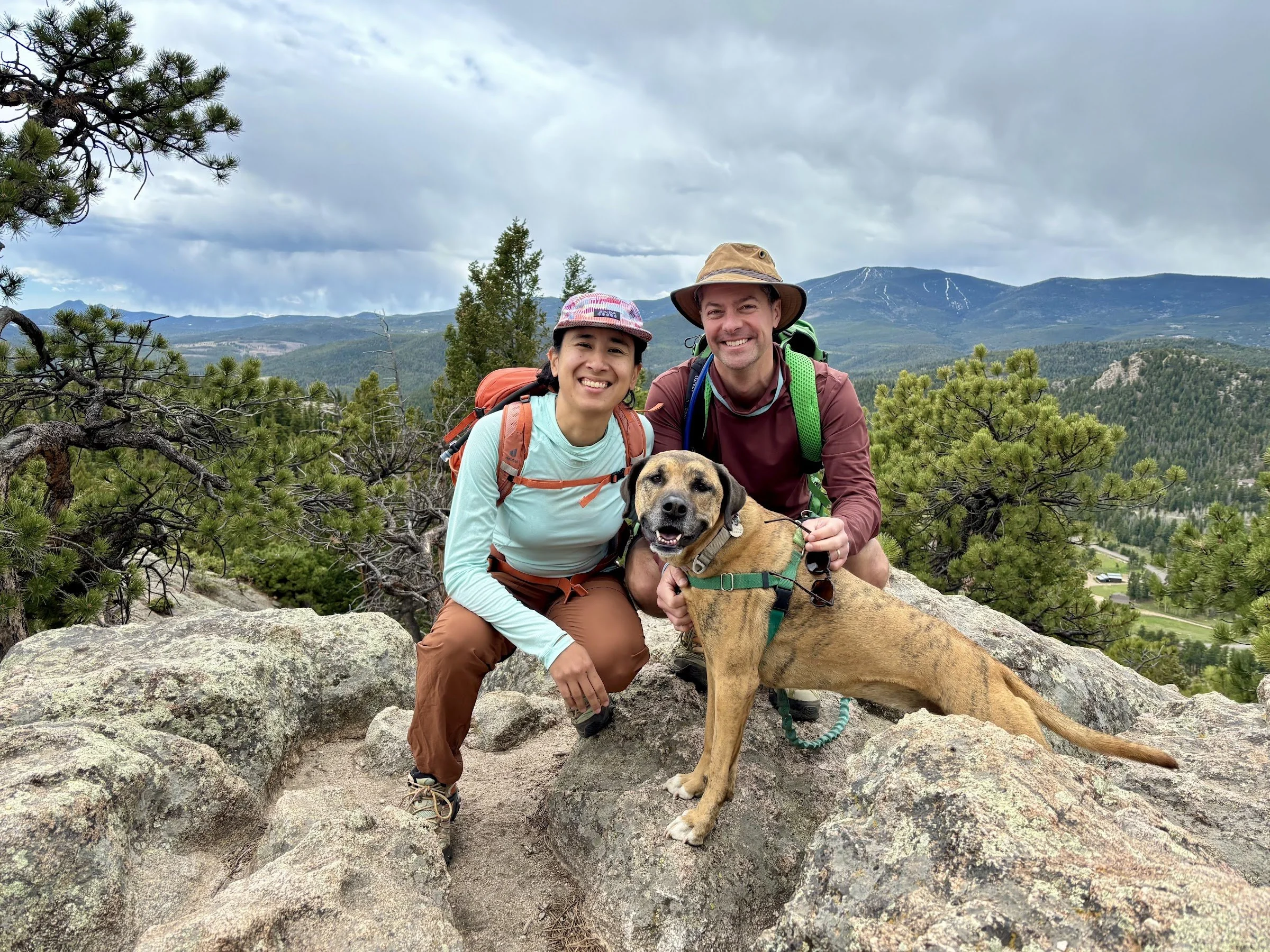 A smiling couple with a backpack and a hat, along with a brown dog, on a rocky mountain trail with a mountain range and trees in the background.