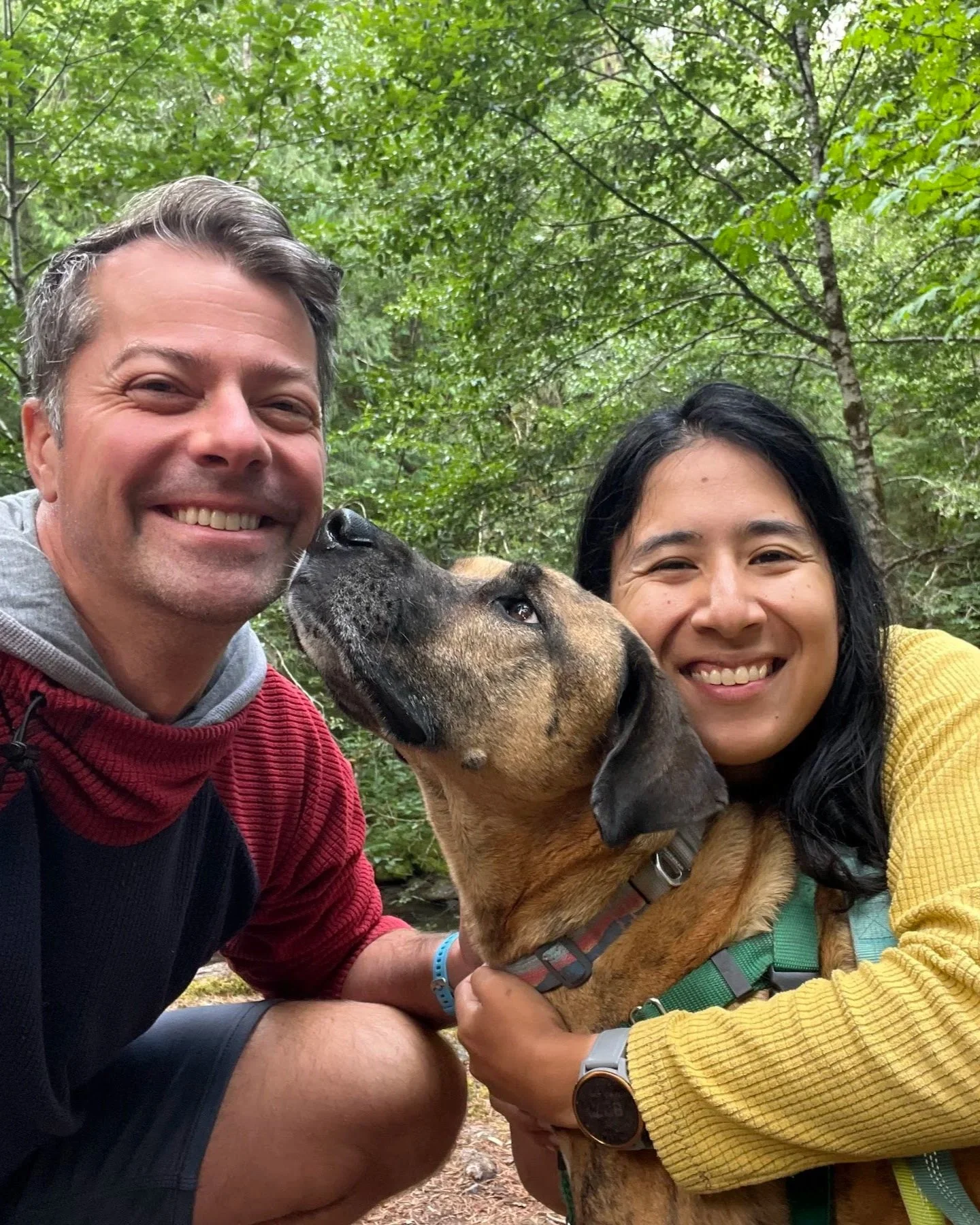 Two smiling people, a man and a woman, are outdoors in a forest with a dog between them. The dog is being kissed on the nose by the man.