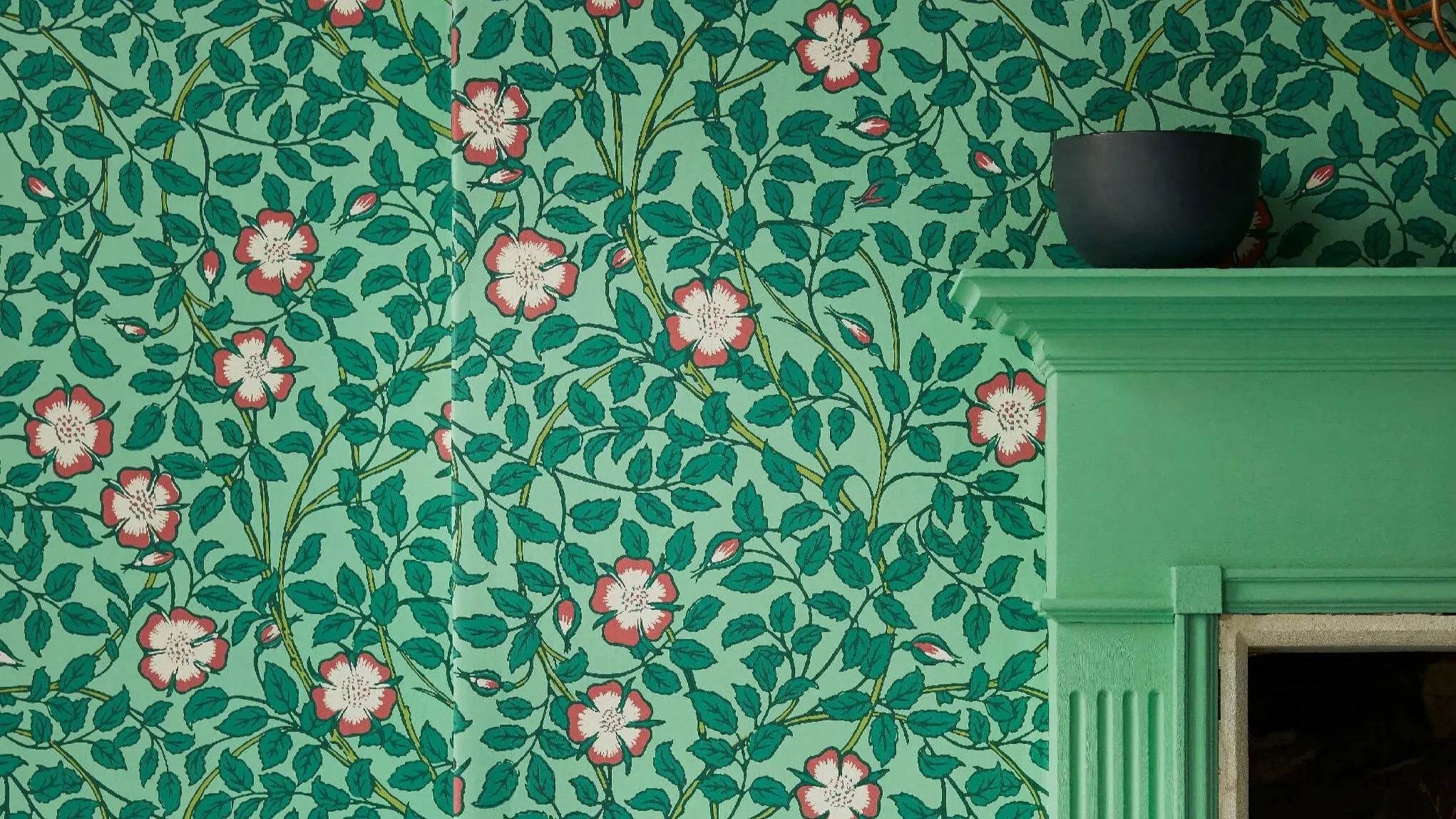 A wall covered with green wallpaper featuring a floral pattern of pink and white flowers with green leaves. Part of a light green fireplace or mantel is visible on the right side of the image.