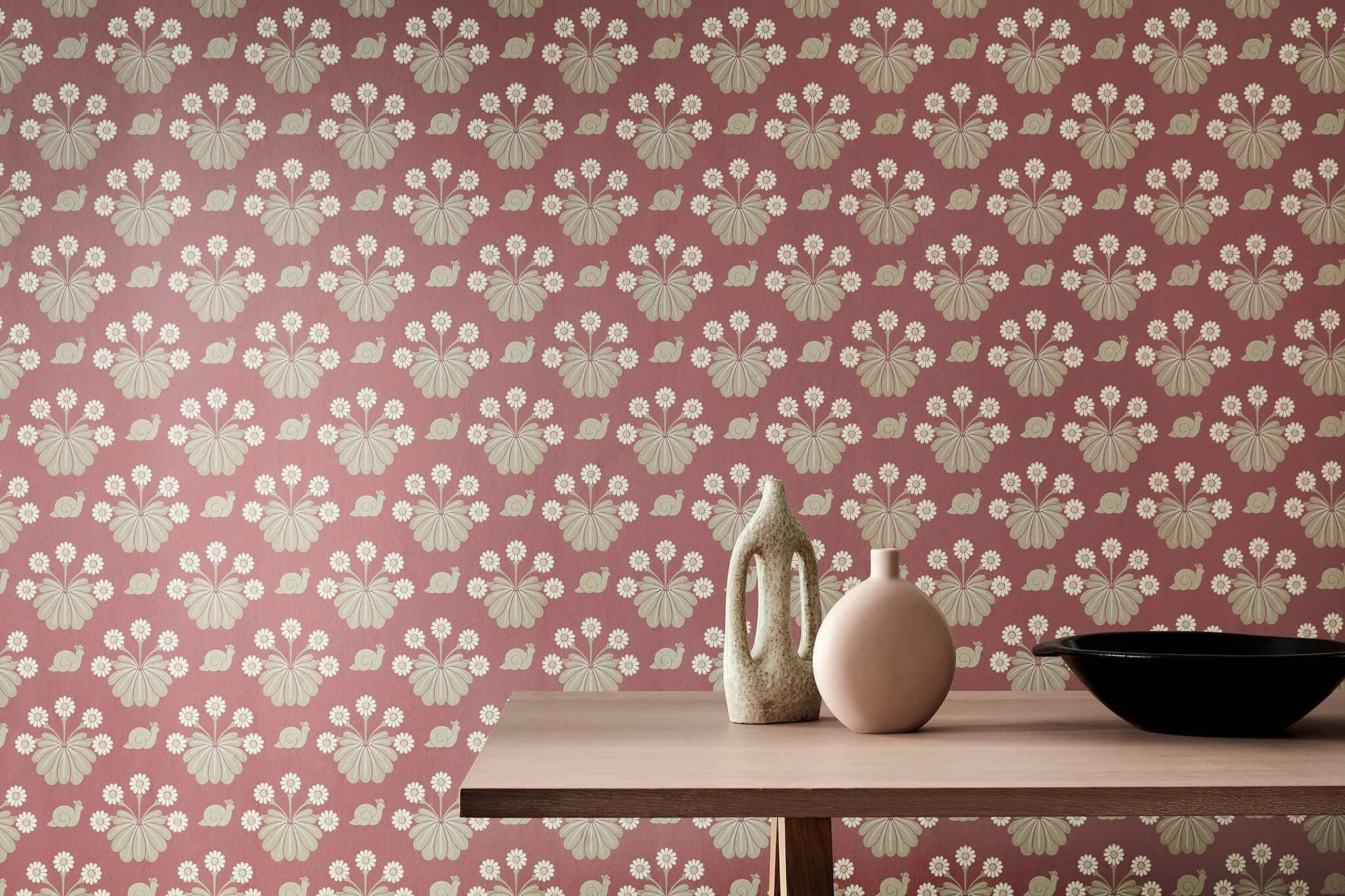 Decorative pink patterned wall with floral and snail motifs, wooden table with three vases of different shapes and colors, and a black bowl.