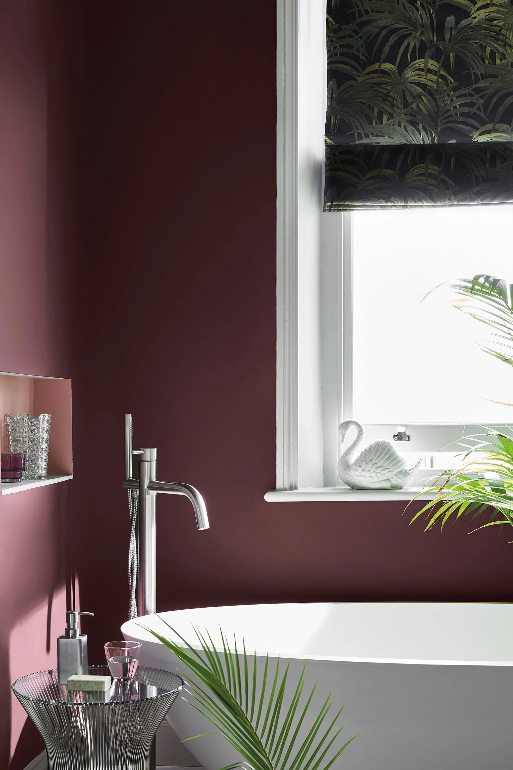 A bathroom with dark mauve walls, a white freestanding bathtub, and a window with tropical-themed roller blind. The windowsill has decorative white swan sculpture and some green plants. There is a chrome floor-mounted bathtub faucet, a round wire storage basket with pink glasses, soap, and toiletries, and a glass of water.
