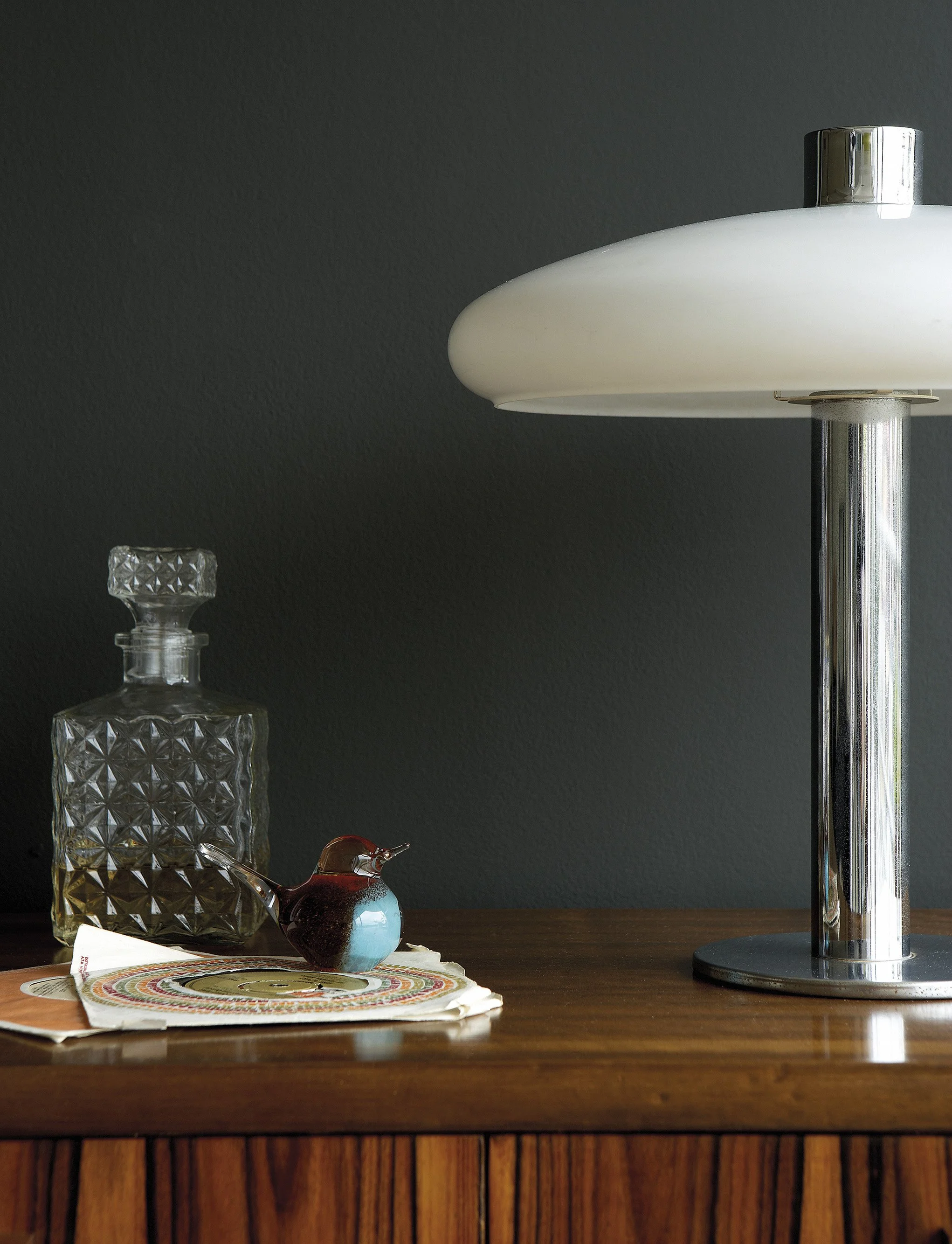 A wooden table with a decorative glass decanter, a small glass bird figurine, a stack of paper plates, and a tall silver lamp against a dark wall.
