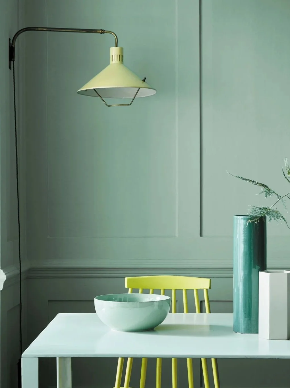 Minimalist interior with a white table, yellow chair, and modern vases on the right, illuminated by a hanging wall-mounted lamp with a white shade.