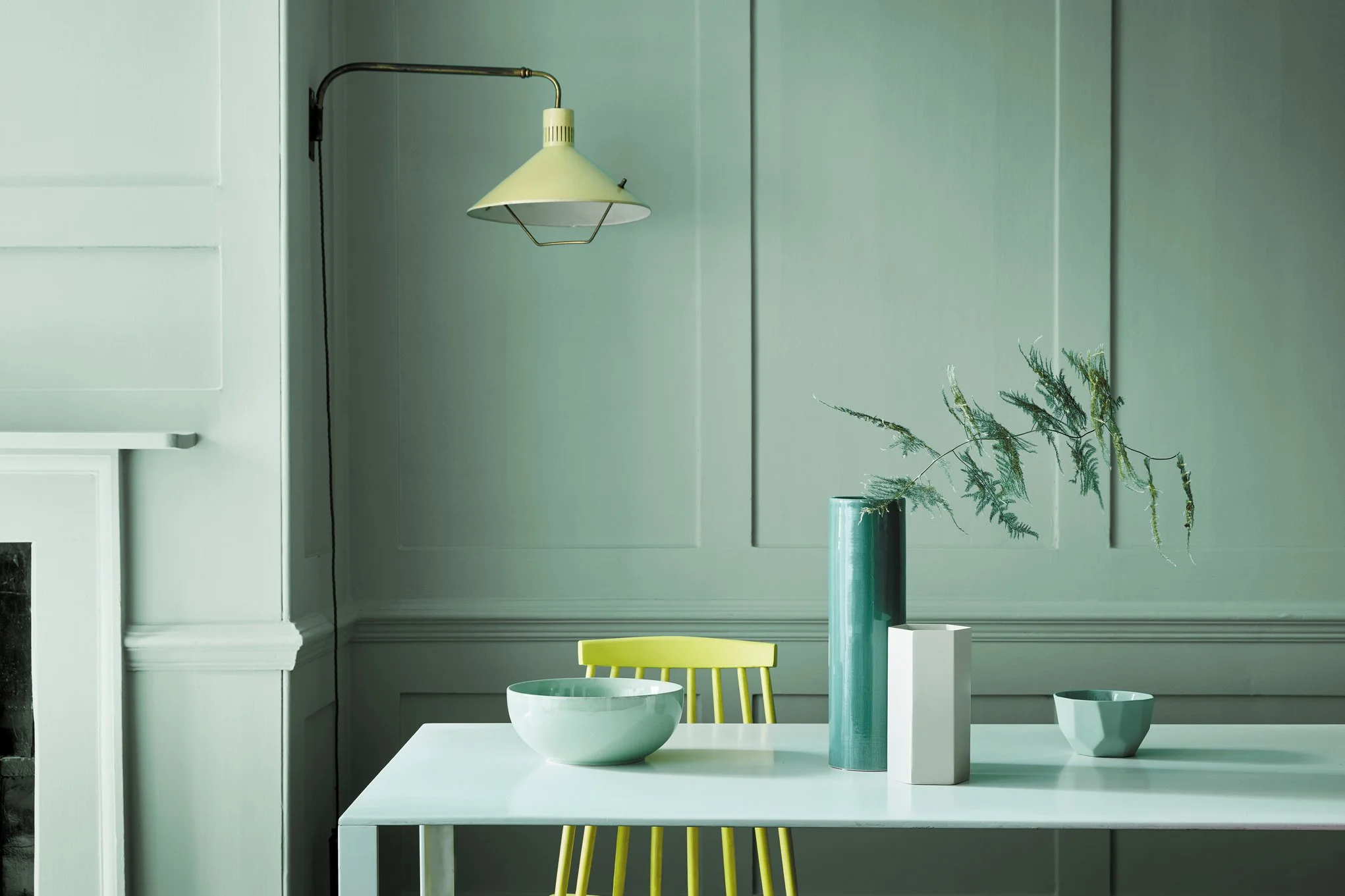 A dining area with a white table, yellow chair, and several ceramic bowls, set against a green wall with paneling and decorative molding.