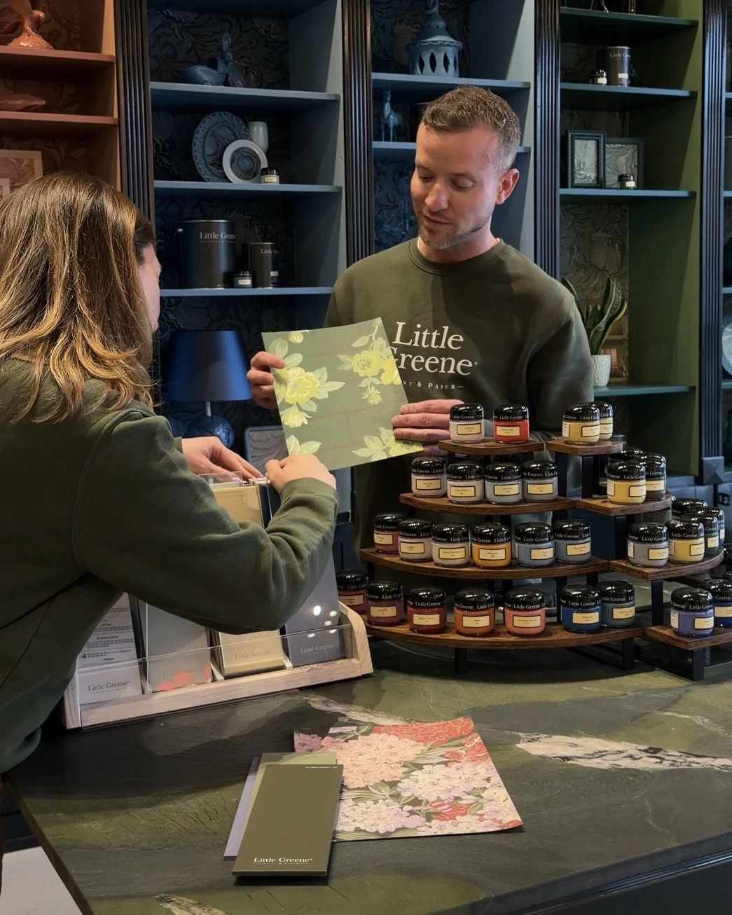 A man and woman are at a store counter, discussing paint color options. The man is holding a sample card with yellow floral patterns and wearing a green sweatshirt with the words 'Little Greene.' Multiple paint jars are stacked on a circular display nearby.