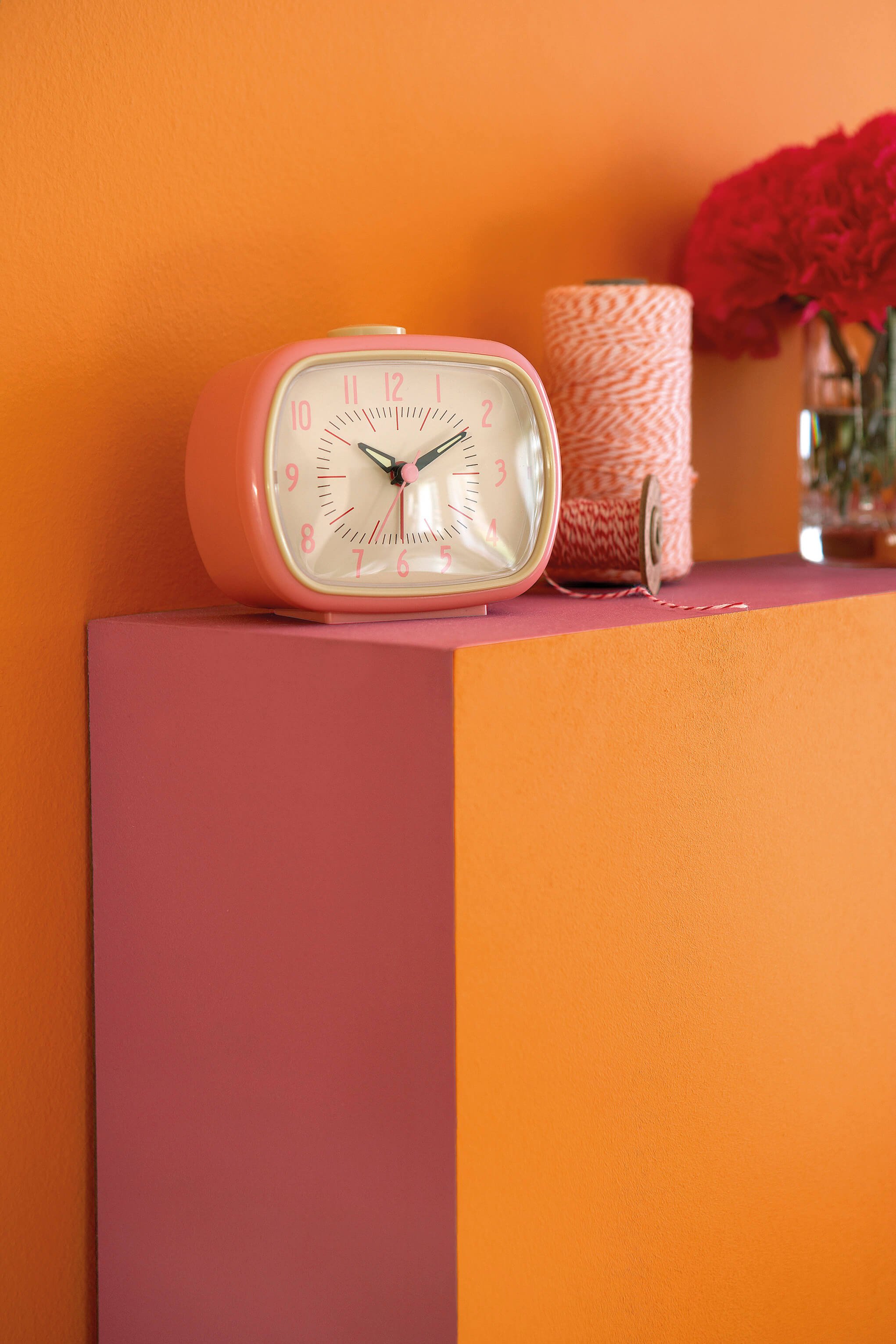 Pink vintage alarm clock with white face on a pink shelf against a bright orange wall, with a spool of orange and white twine and a glass vase with red flowers in the background.