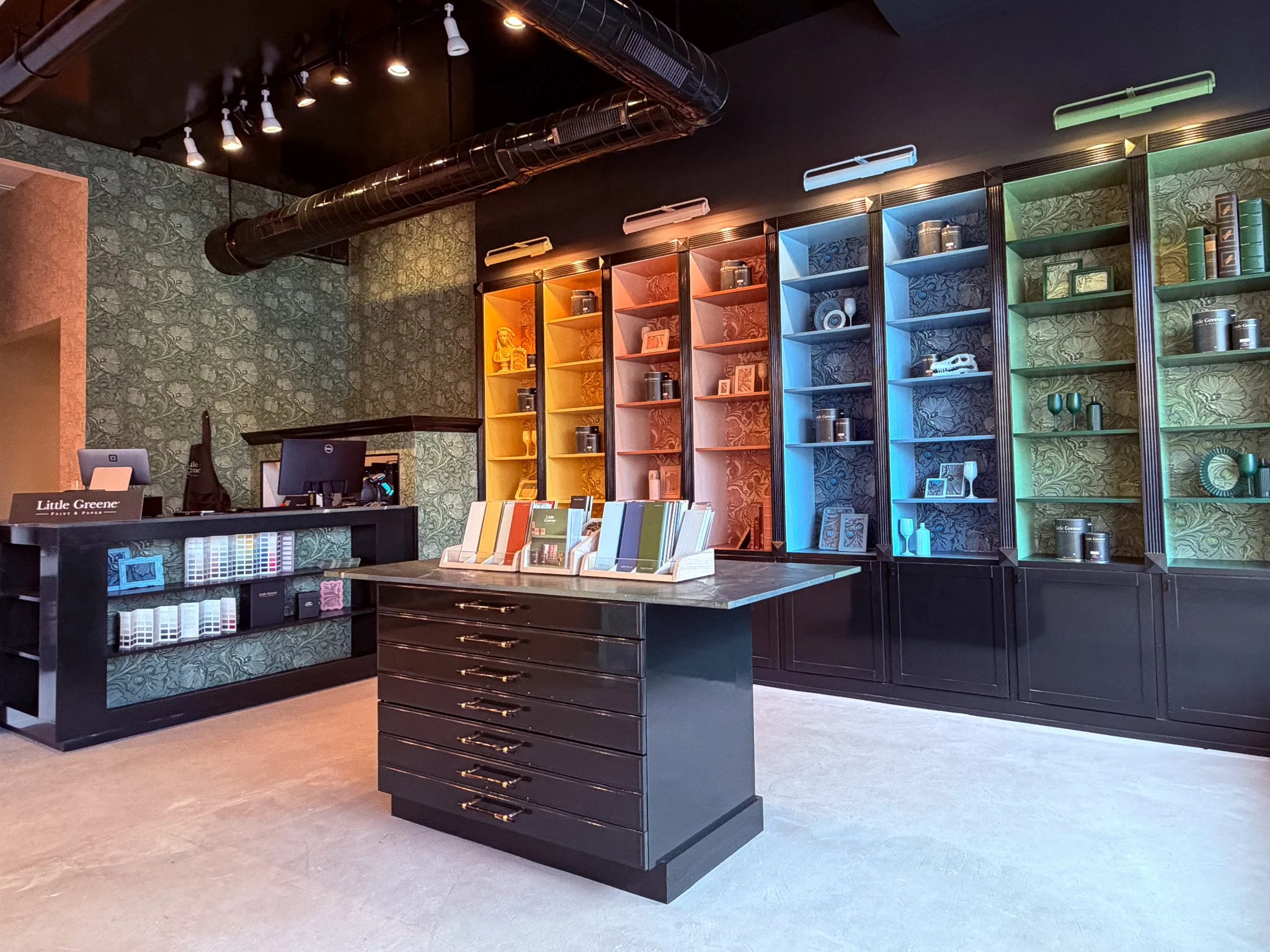An interior of a store with dark wooden shelves filled with books and decorative items, a black central island with drawers, colorful books, and artwork, and a wall with leaf-patterned wallpaper and colorful lighting fixtures.