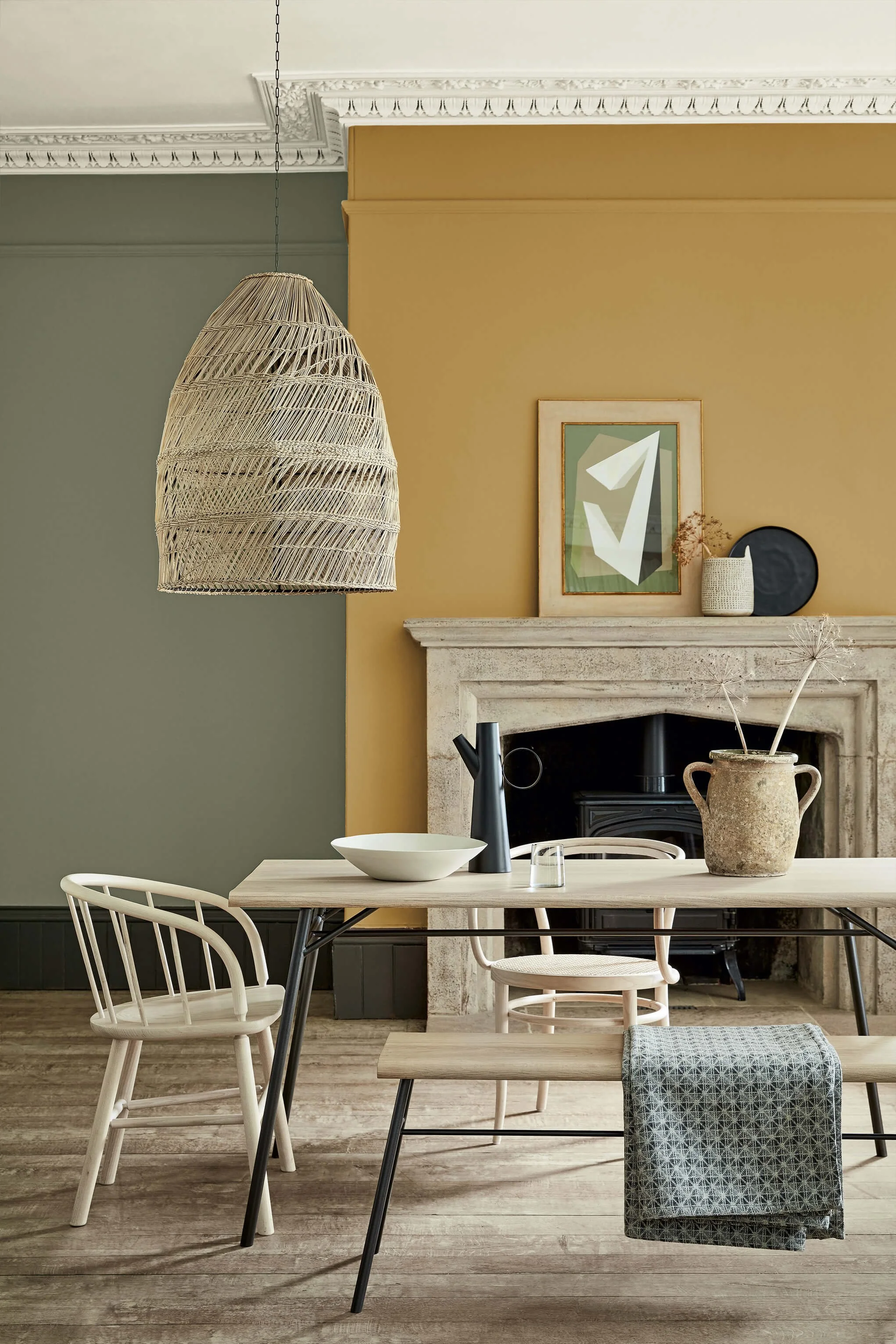 A modern dining room with a wooden table, white chairs, and a fireplace. Decor includes a large woven hanging lamp, framed artwork, a vase, and decorative items on the mantel, with color walls in green and mustard yellow.