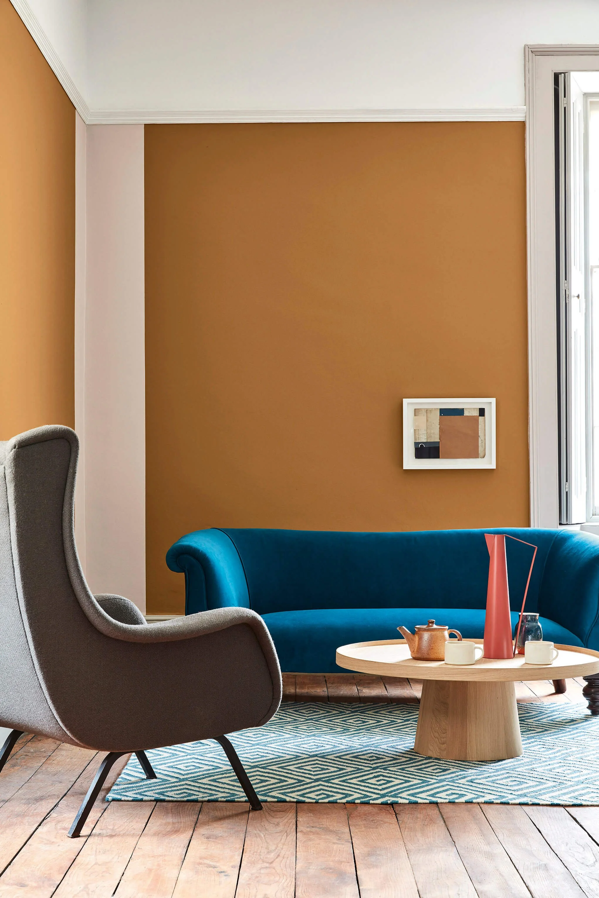 Living room with a gray armchair, a teal loveseat, a wooden coffee table with a teapot and cups, a patterned rug, a gold and white wall, a framed artwork, and a window.