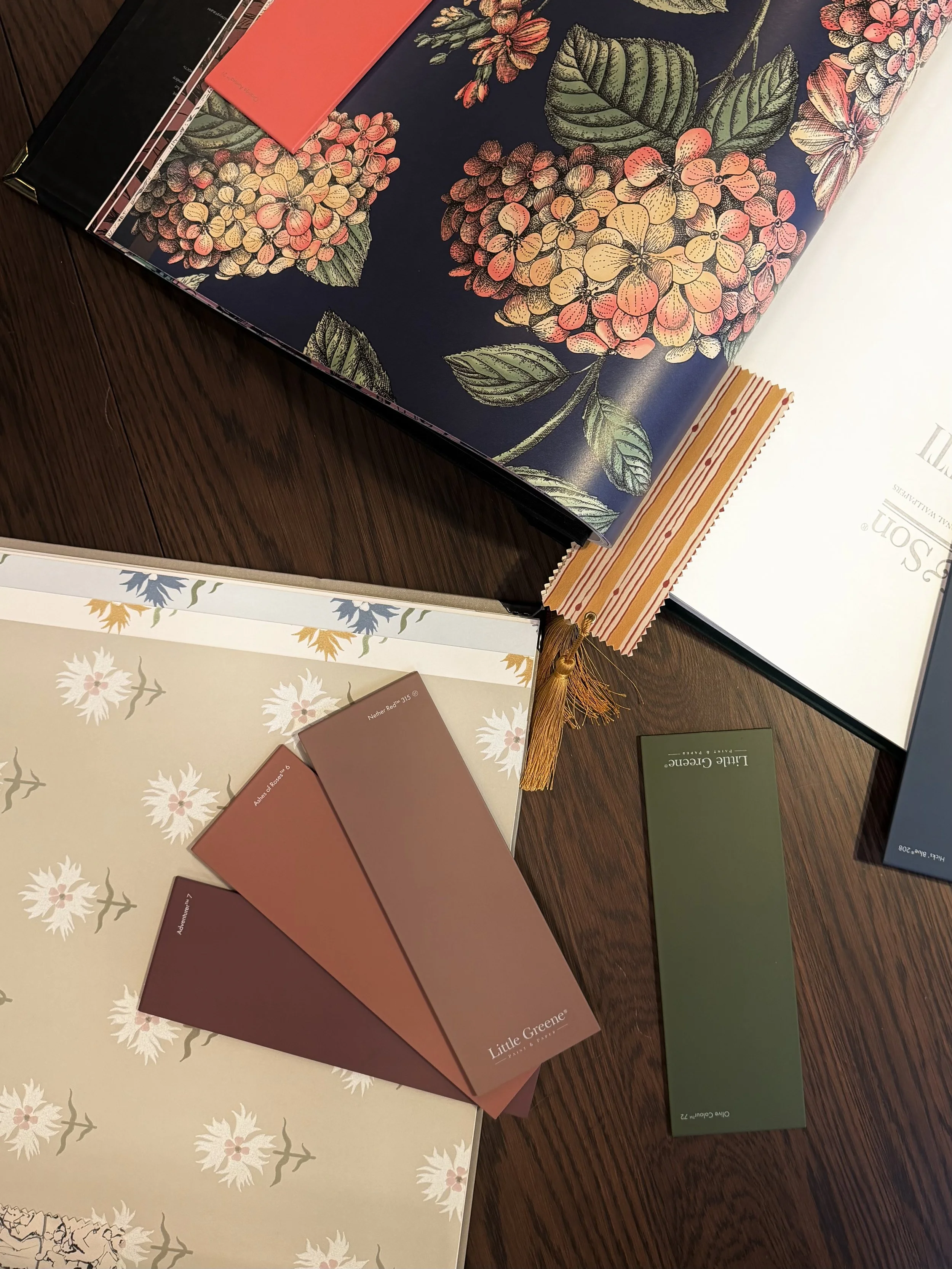 Sample color swatches, books, and fabric swatches on a wooden surface, with a floral patterned fabric and decorative tassel.