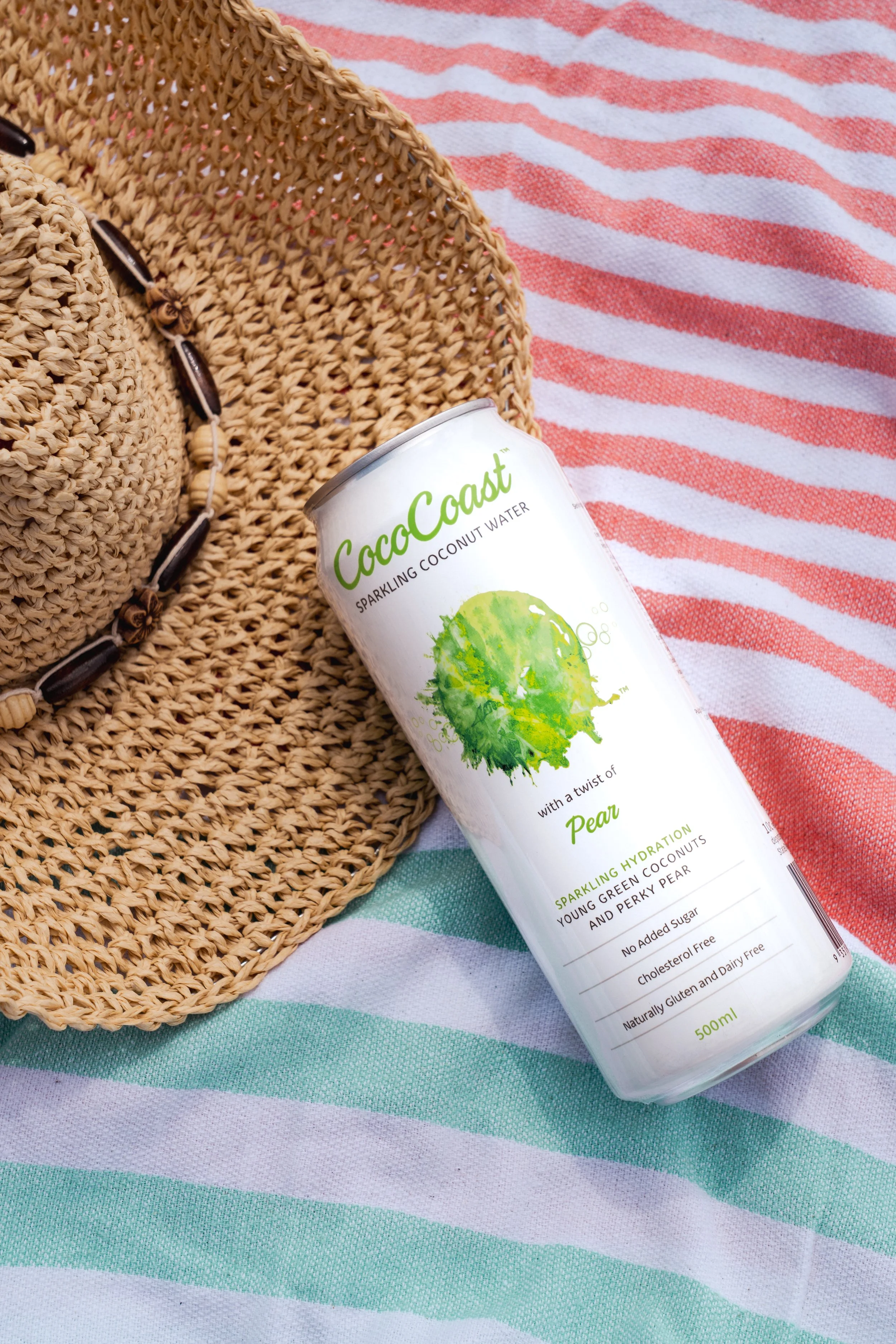 A can of CocoCoast sparkling coconut water with a twist of pear, lying on a striped beach towel next to a woven straw hat.