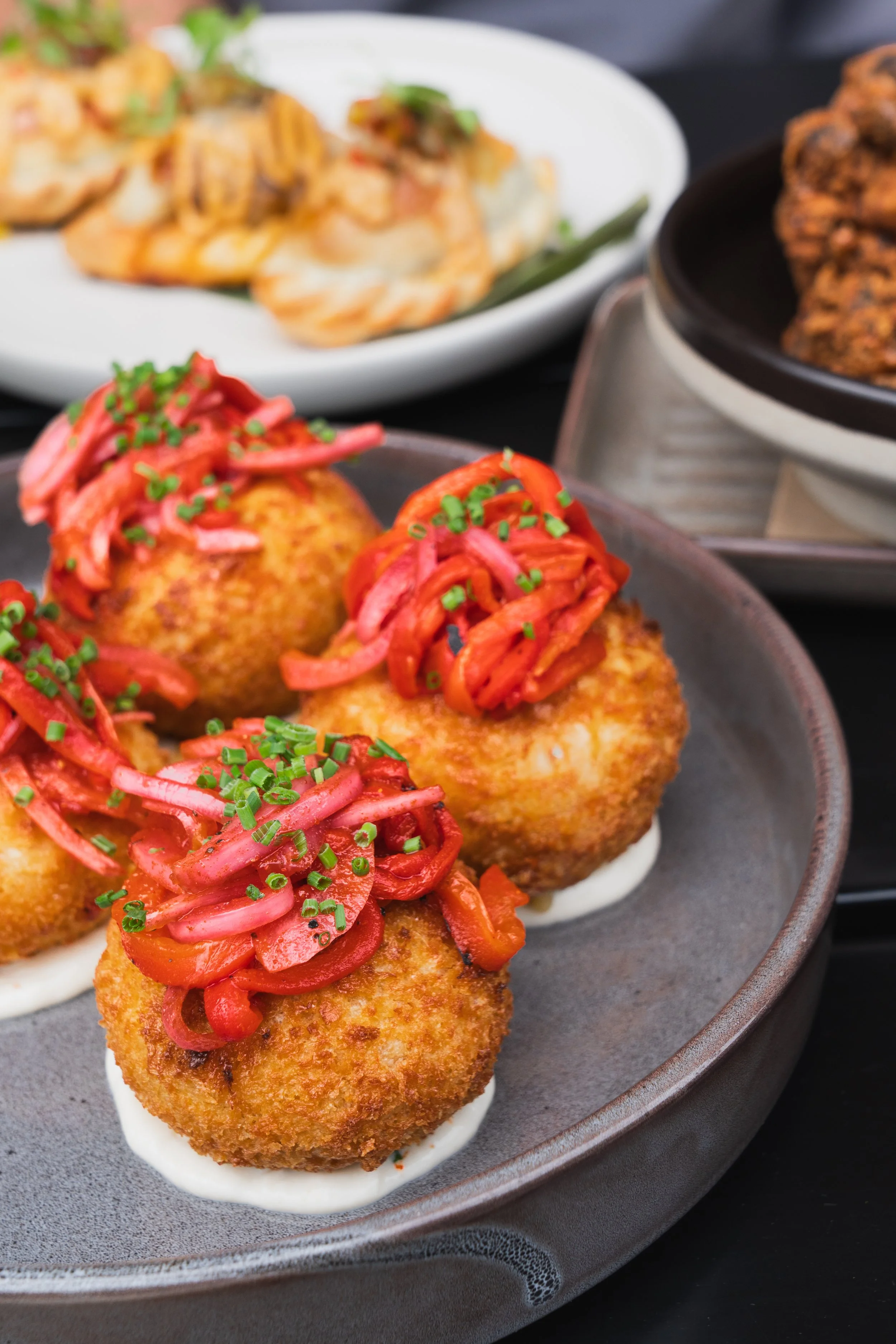 Close-up of three croquettes topped with pickled vegetables and chopped chives on a dark plate with other dishes in the background.