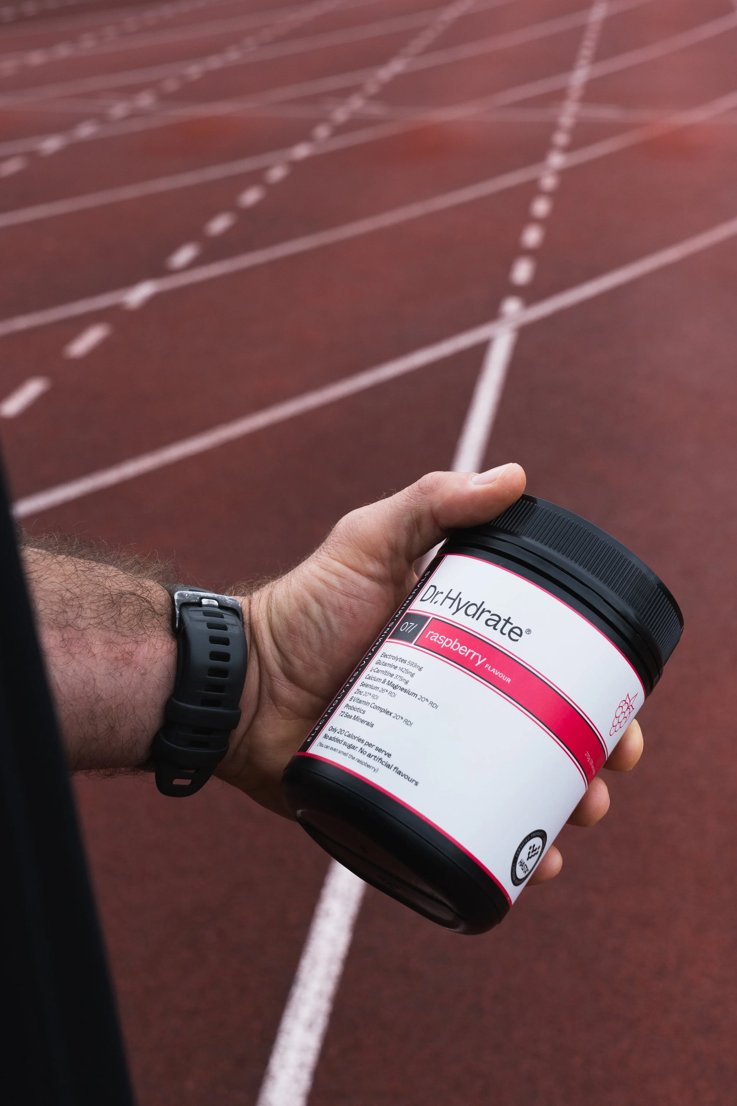 A person holding a container of Dr. Hydrate raspberry flavored electrolyte powder on a red running track.