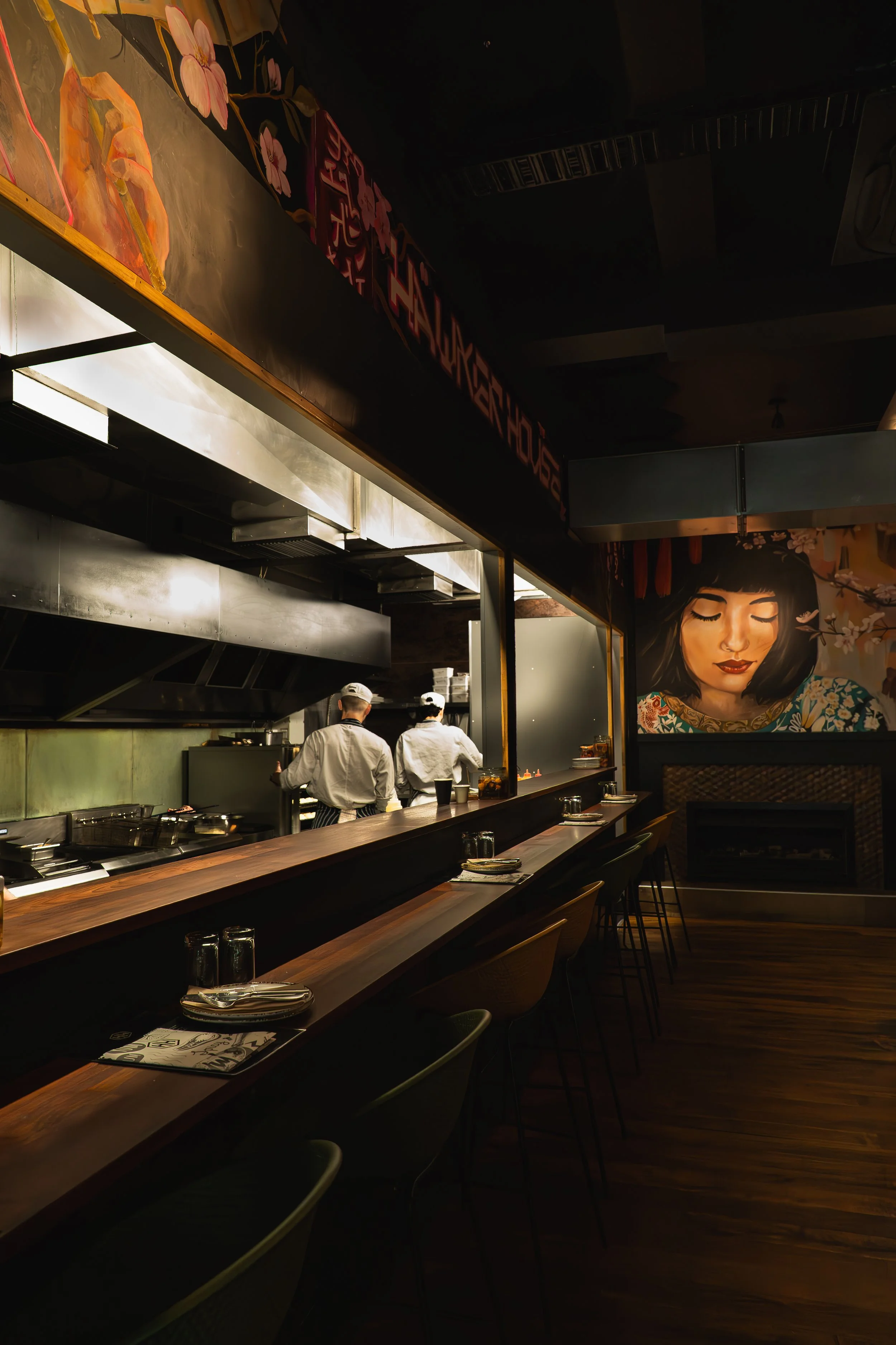 Inside a modern restaurant with a bar counter, where two chefs are working in the open kitchen. The decor includes large colorful murals of a woman's face and floral art on the walls.