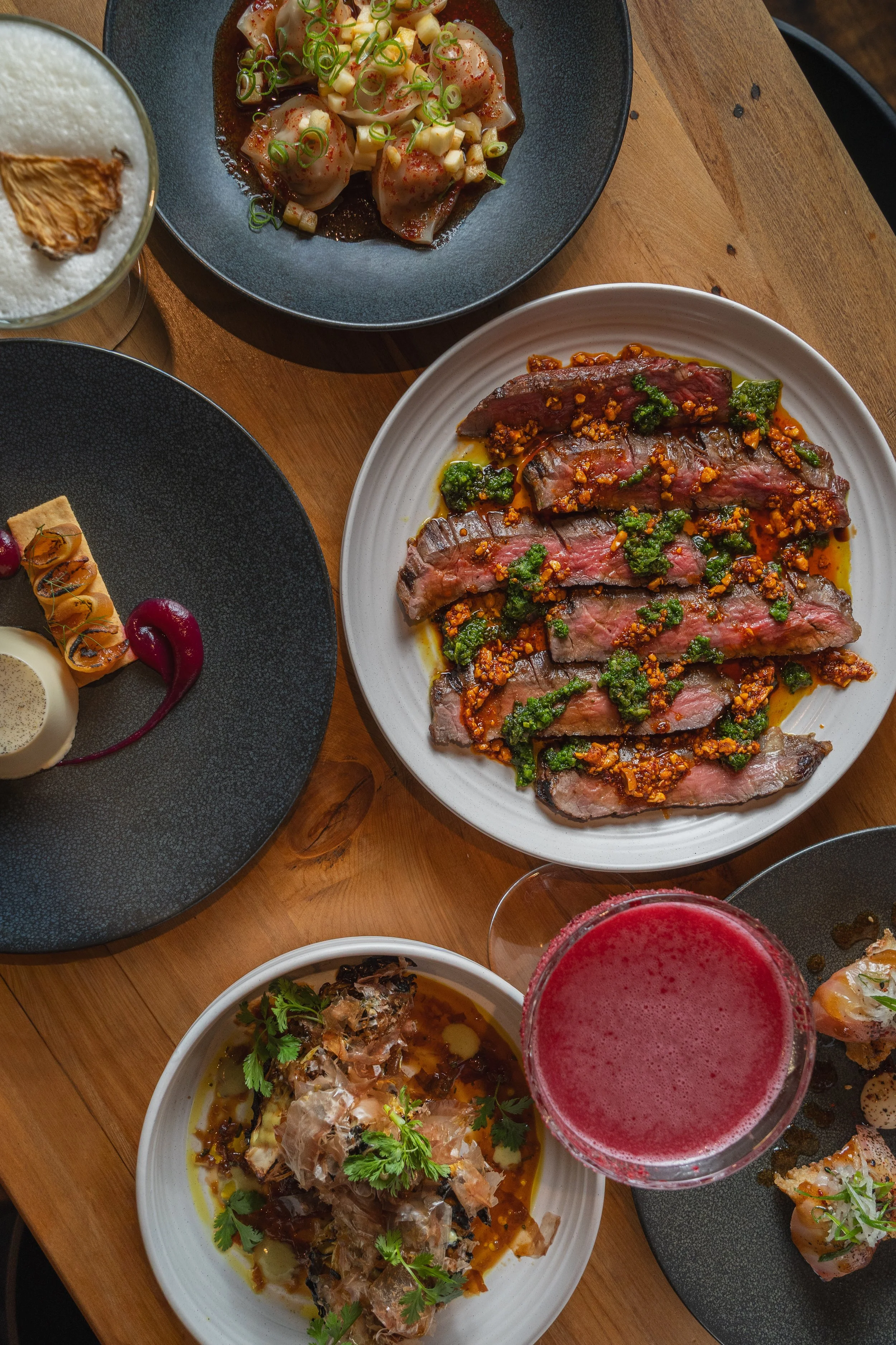 A variety of dishes on a wooden table, including sliced beef with herbs and sauce, a plate of sushi rolls, a bowl of oysters topped with herbs, a dessert with a red sauce, and a cocktail with foam and a dried fruit garnish.