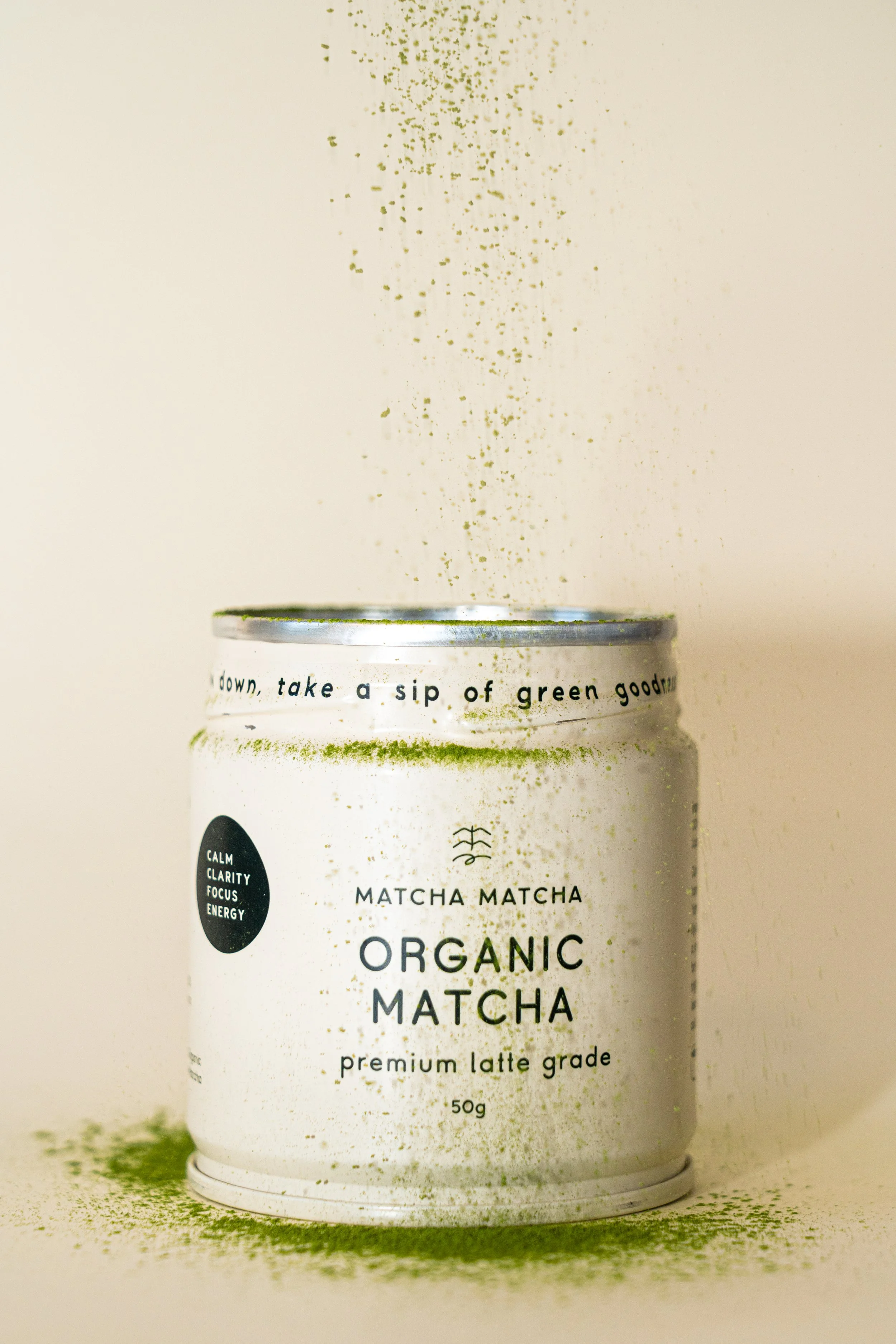 Open can of organic matcha latte powder with green matcha powder being sprinkled above it, creating a scattering effect on the surface and around the can.