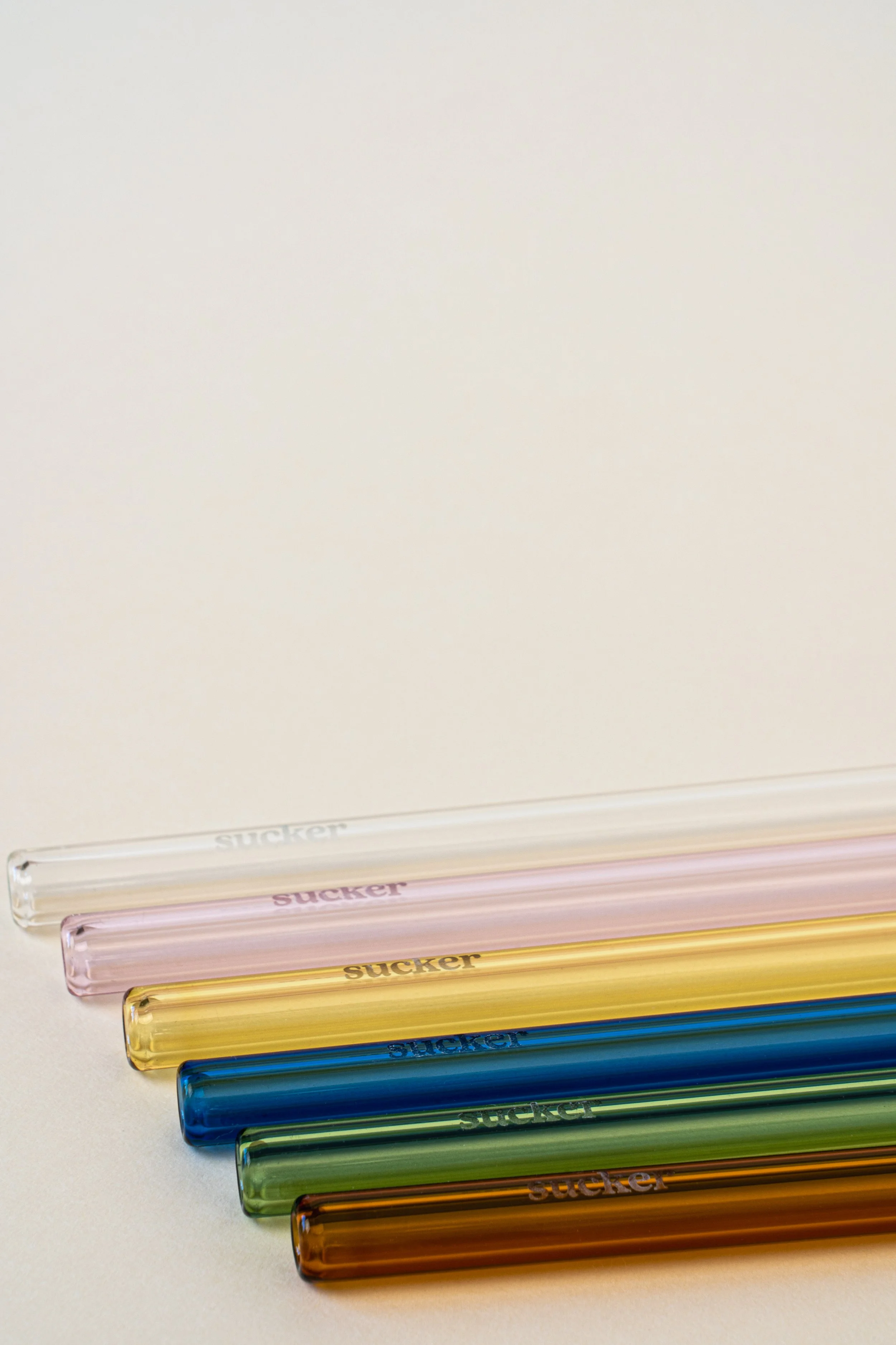 Six colorful reusable glass drinking straws in clear, pink, yellow, blue, green, and amber, each with the word 'sucker' printed on them, arranged diagonally on a white surface.
