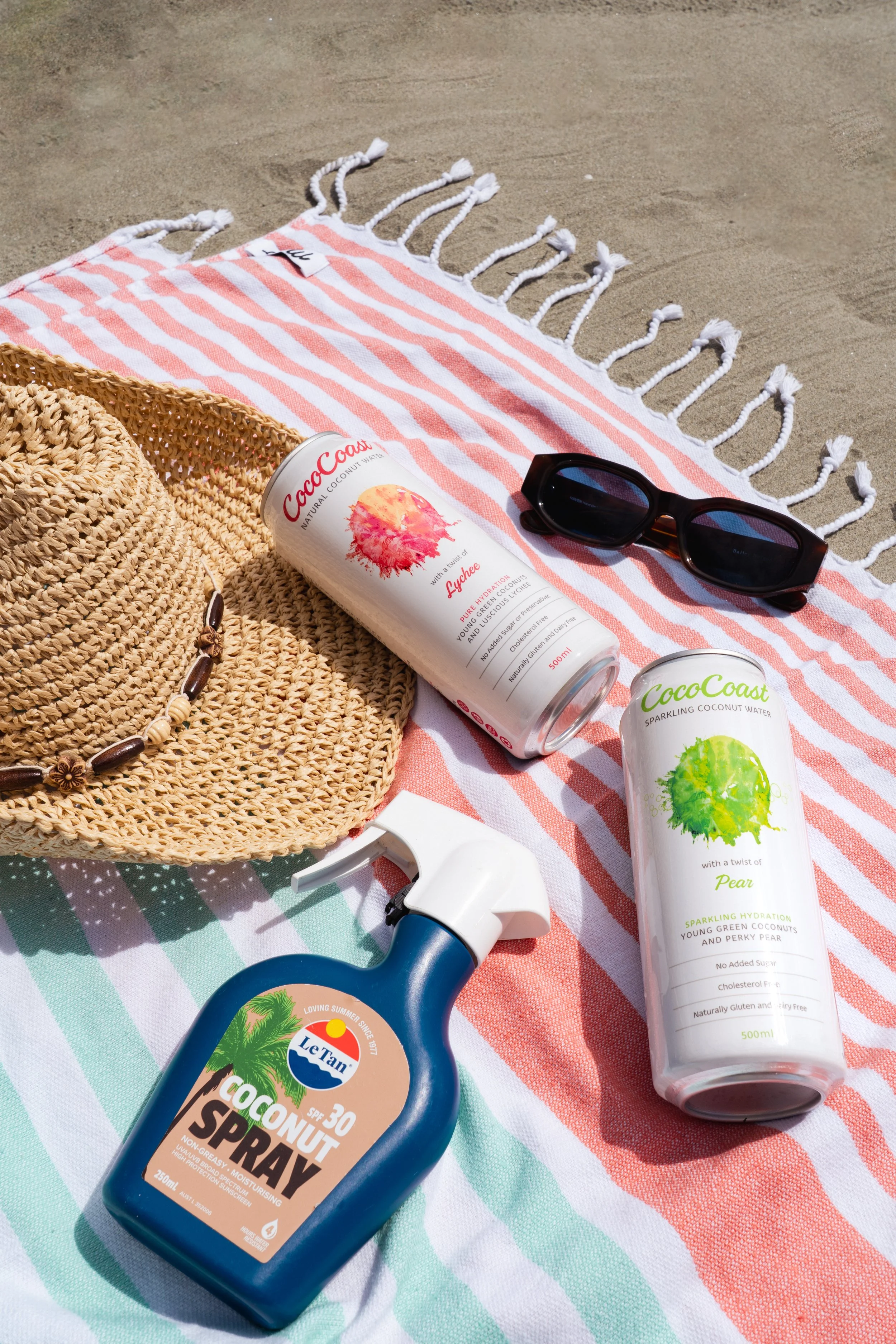 Beach towel with a striped pink and white pattern, a straw sun hat with a shell necklace, a pair of sunglasses, and three coconut water cans on the sand, with one labeled Lychee, one labeled Pear, and a coconut water spray bottle.