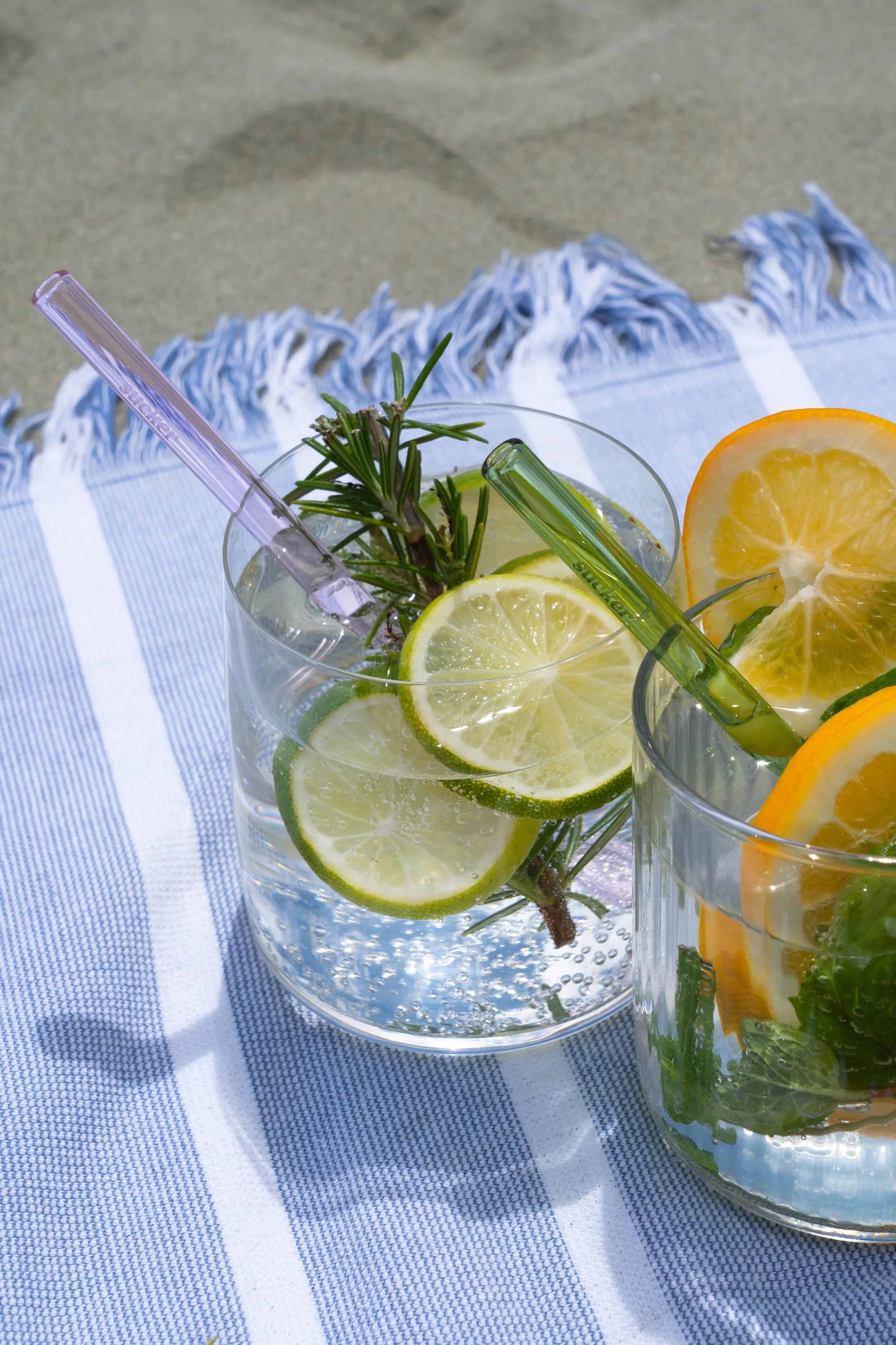 Reusable glass straws sitting in two glasses of infused water with lemon, lime, and herbs, placed on a blue and white striped cloth.