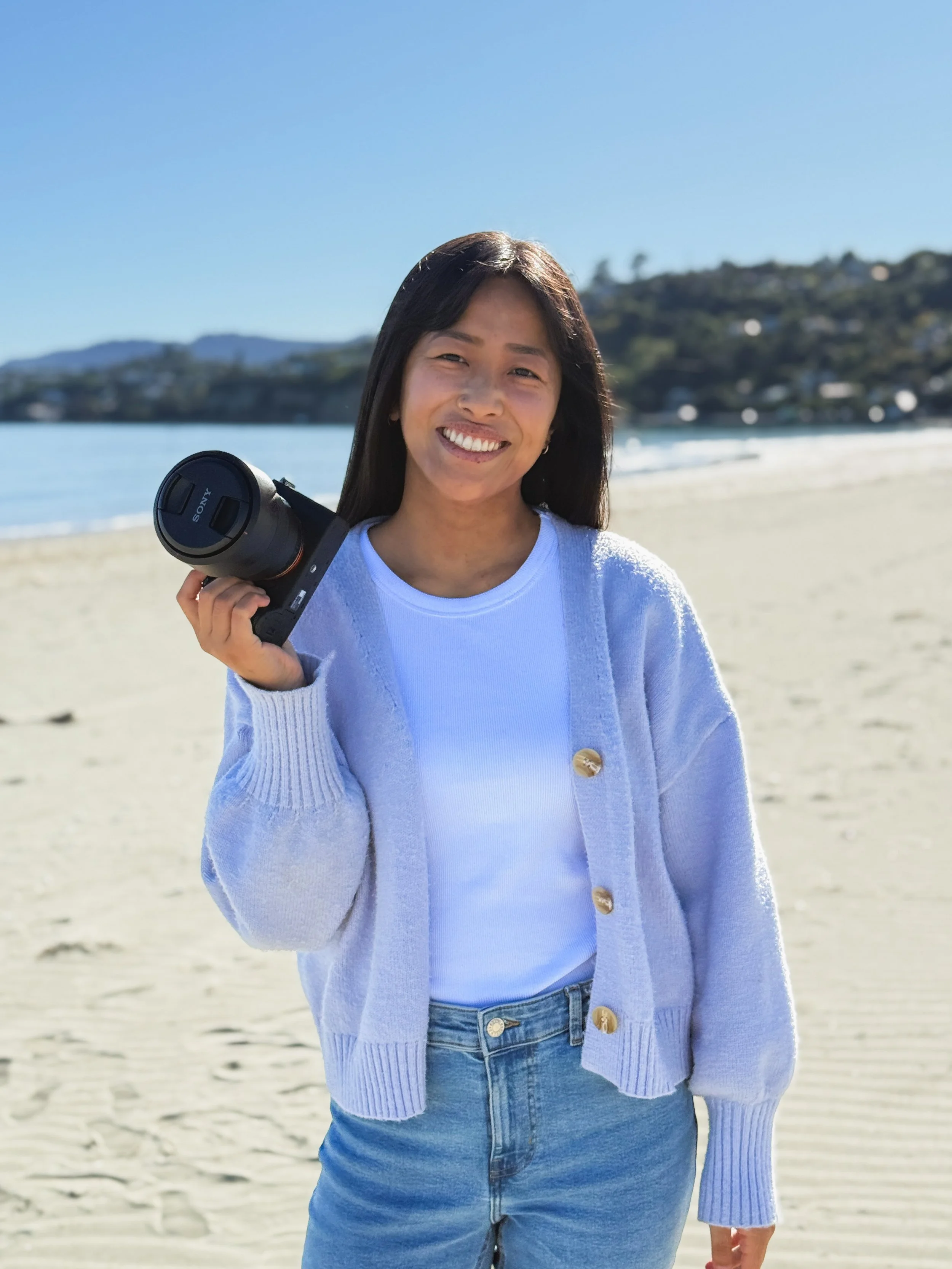 Smiling woman holding a camera on a sunny beach with sand and ocean in the background
