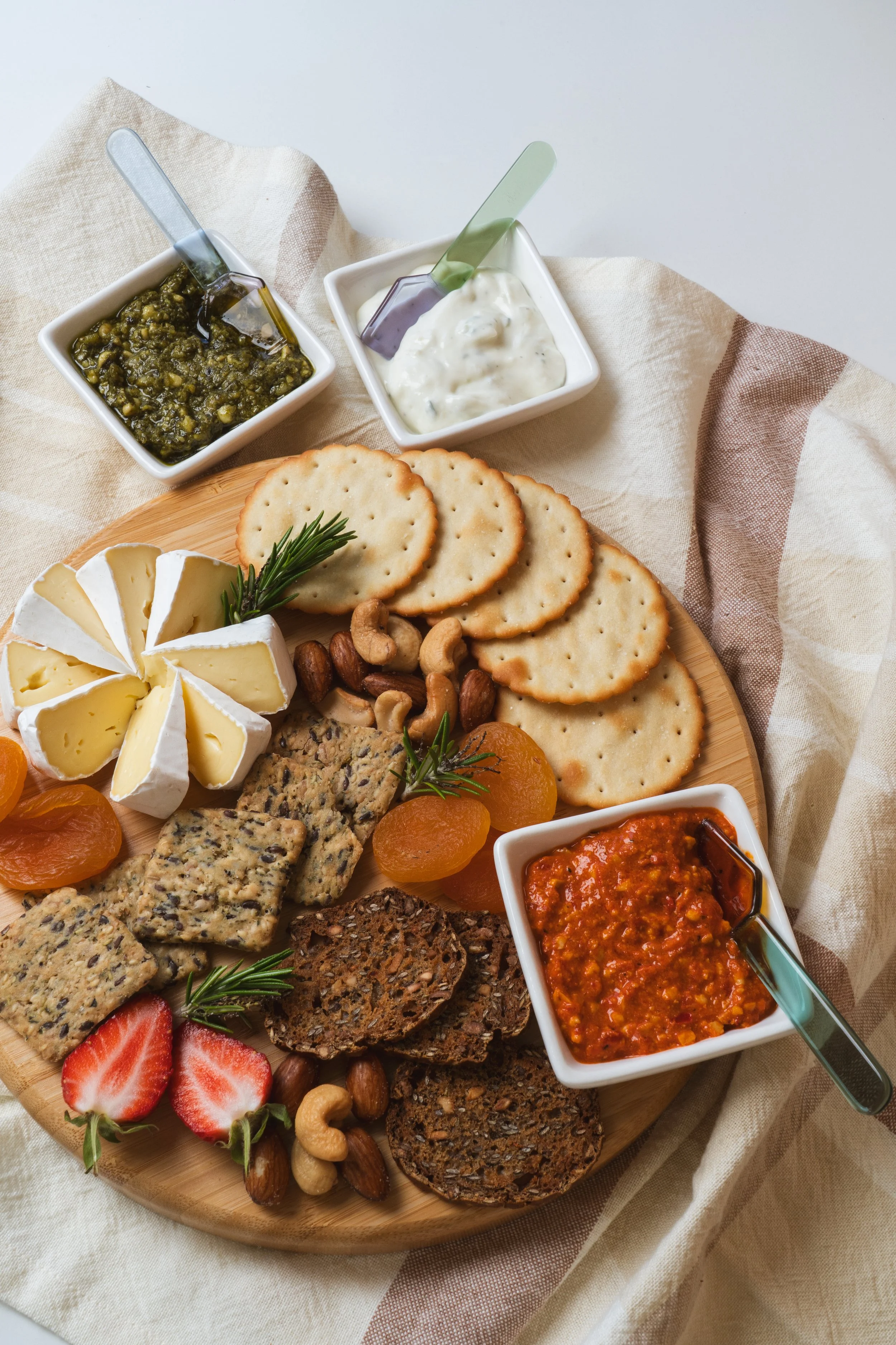 A cheese board with crackers, sliced brie, dried apricots, strawberries, mixed nuts, seed crackers, and spreads of chutney, yogurt-based dip, and spicy tomato relish.