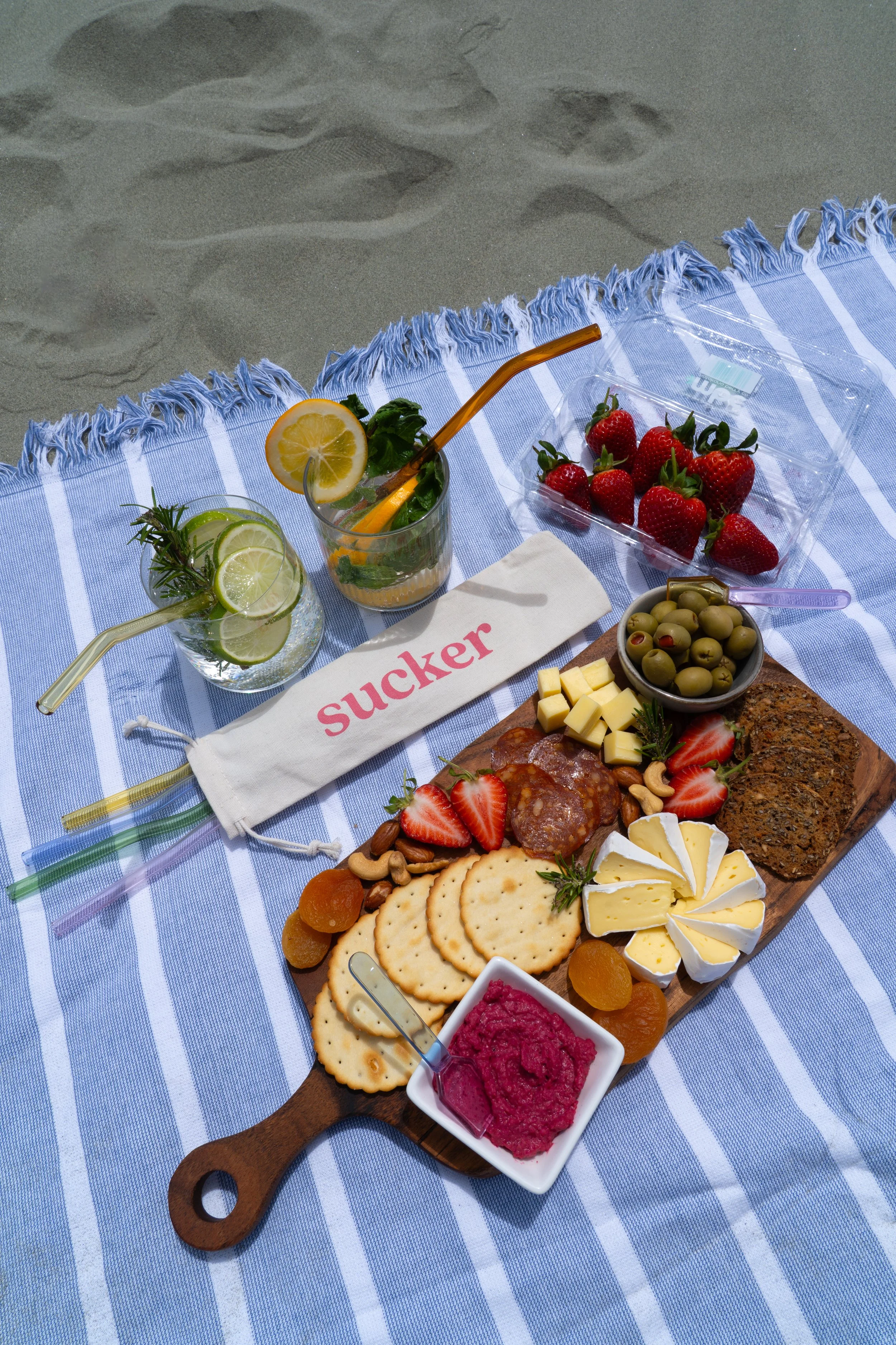 A picnic spread on a blue and white striped blanket at the beach, featuring cheese, strawberries, olives, crackers, and drinks with lemon and lime slices