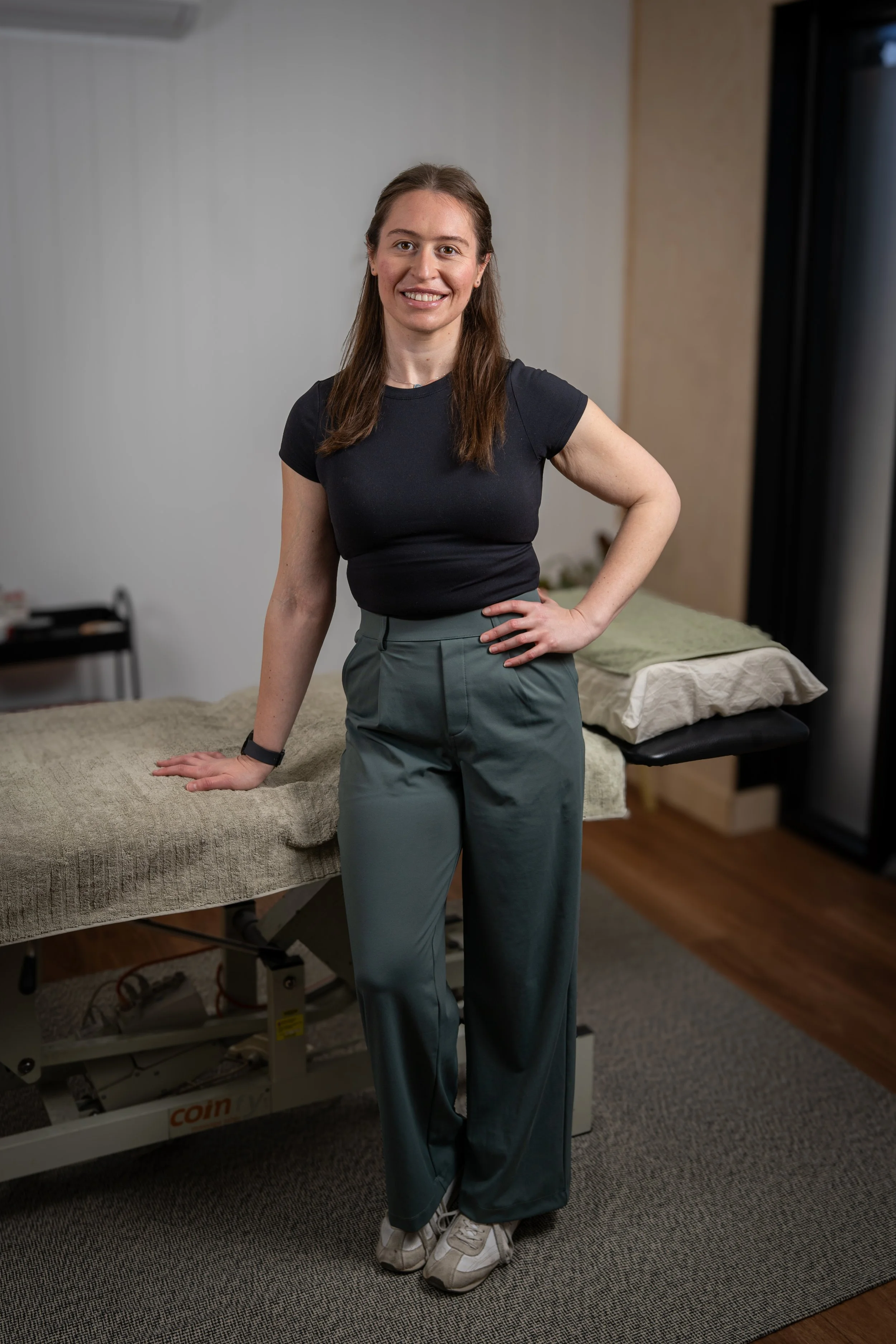 A woman stands next to a massage table in a room. She is wearing a black T-shirt, loose gray pants, and sneakers. She has long, brown hair and is smiling at the camera. The room has a bed with pillows, a curtain, and a small side table.