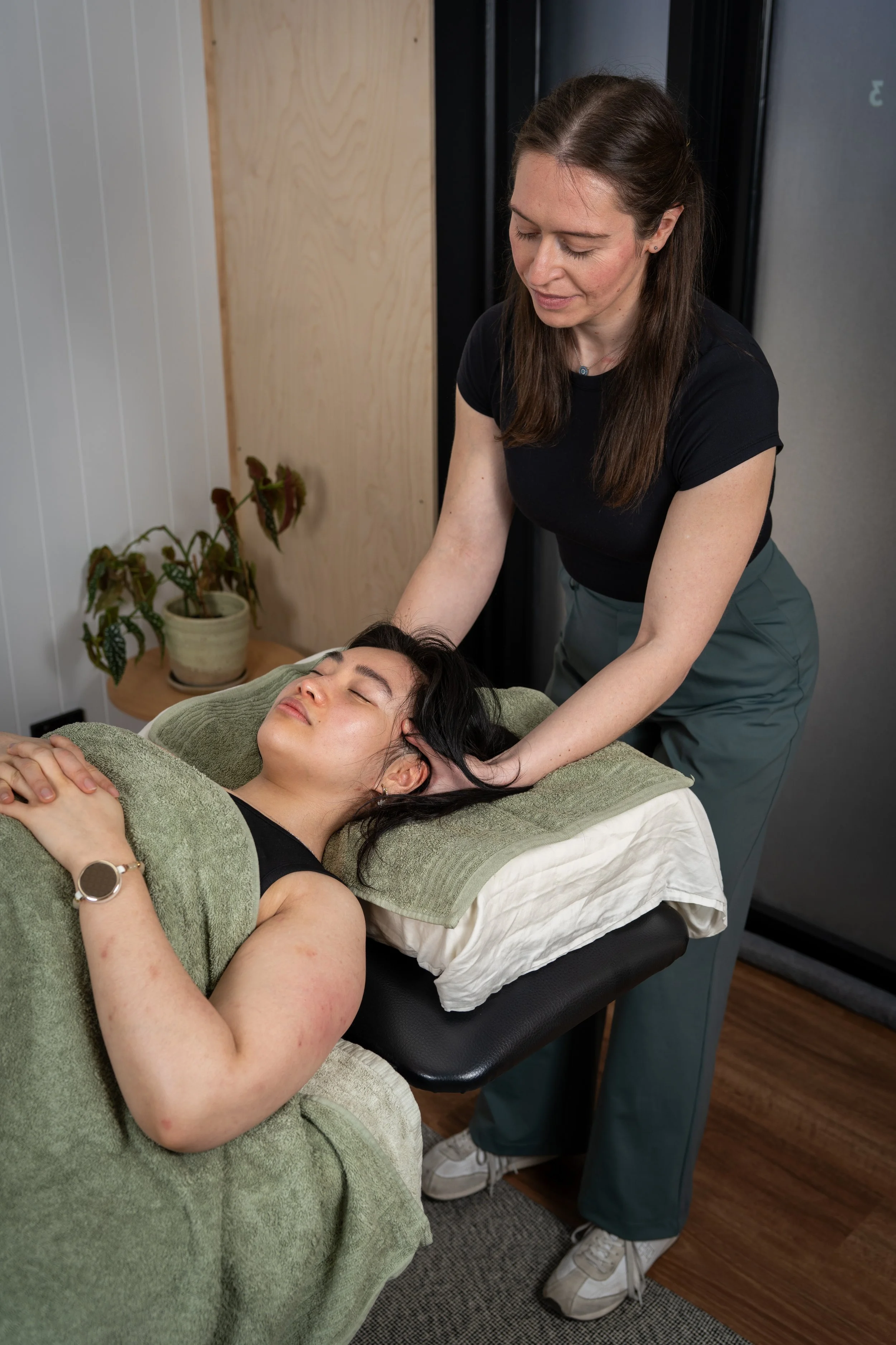 A woman with dark hair and a green watch is lying on a massage table with her eyes closed, covered with a green towel. Another woman with long brown hair is standing next to her, performing a massage or therapy with her hands on the client's head and neck. There is a potted plant in the background on a wooden surface.