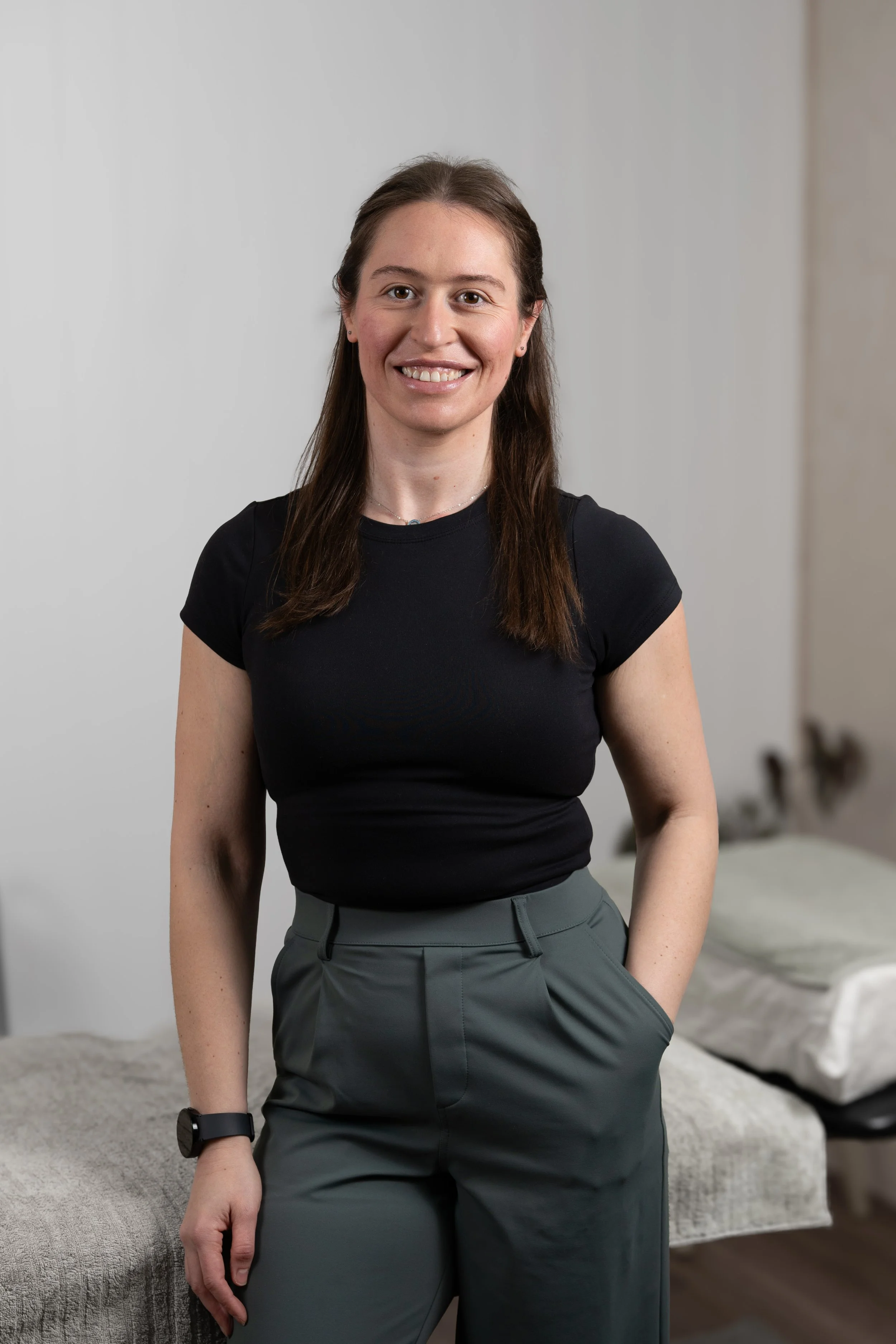 A woman Osteopath/Allied health practitioner with long brown hair, smiling, wearing a black t-shirt and green high-waisted pants standing in a modern interior with a white and beige wall background.