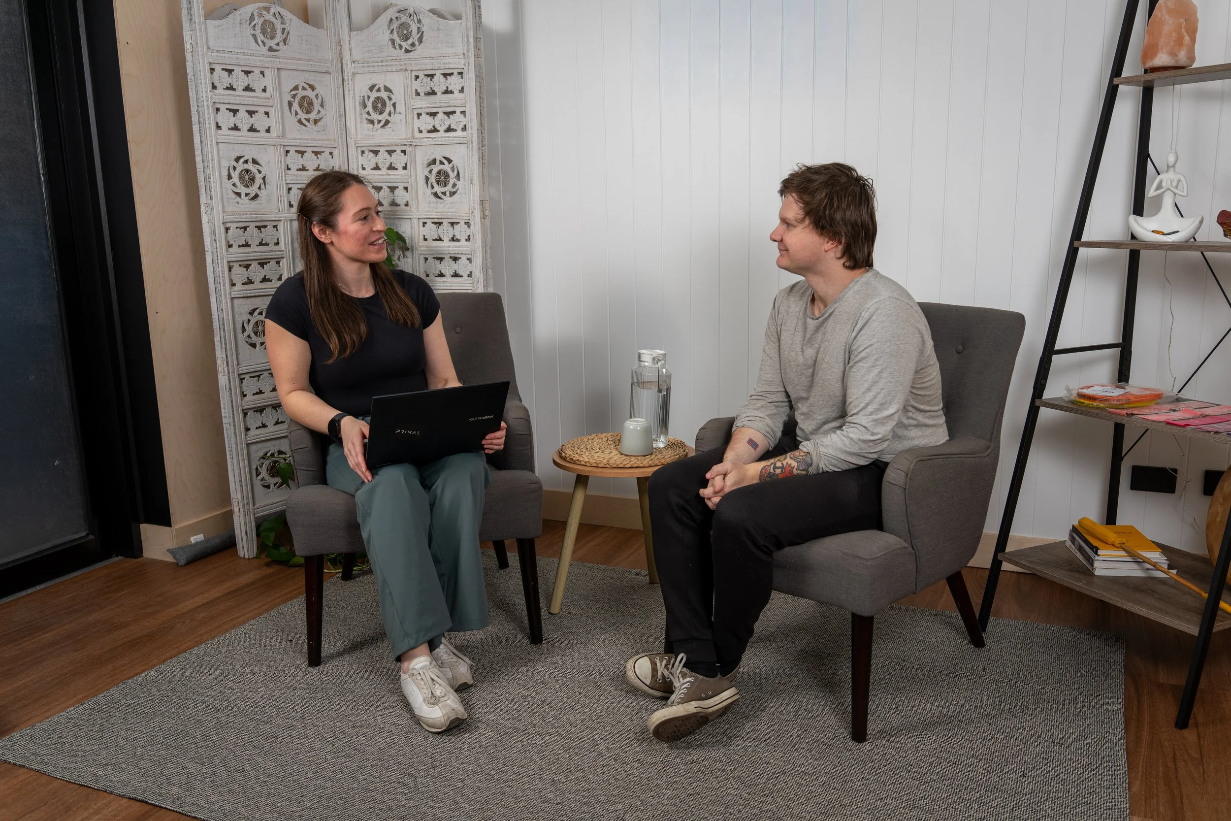 Two people, an Osteopath and her client/patient sitting on gray chairs in a cozy clinic room, engaged in conversation. The Osteopath is taking the client's history and gathering information regarding the present pain/injury/movement complaint.