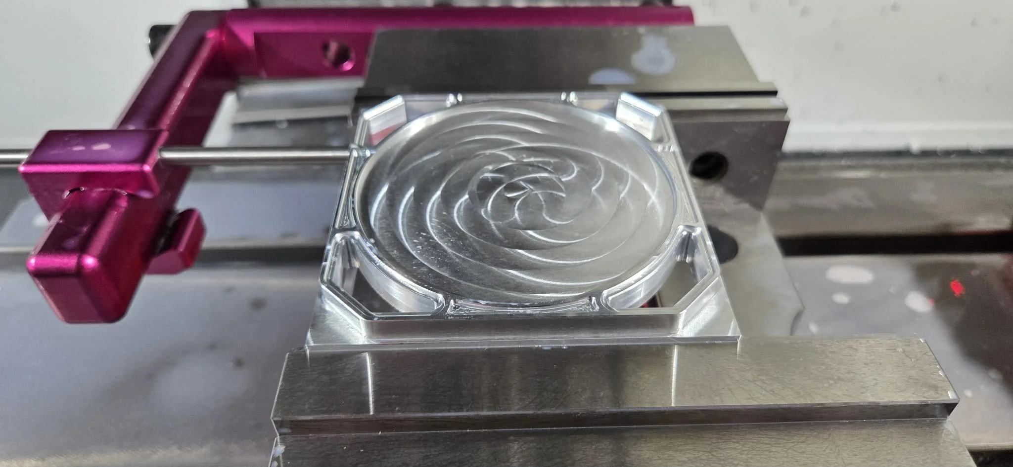 A laboratory setup with a metallic platform holding a transparent, circular component with spiral grooves, secured by a pink clamp on a metal rail.