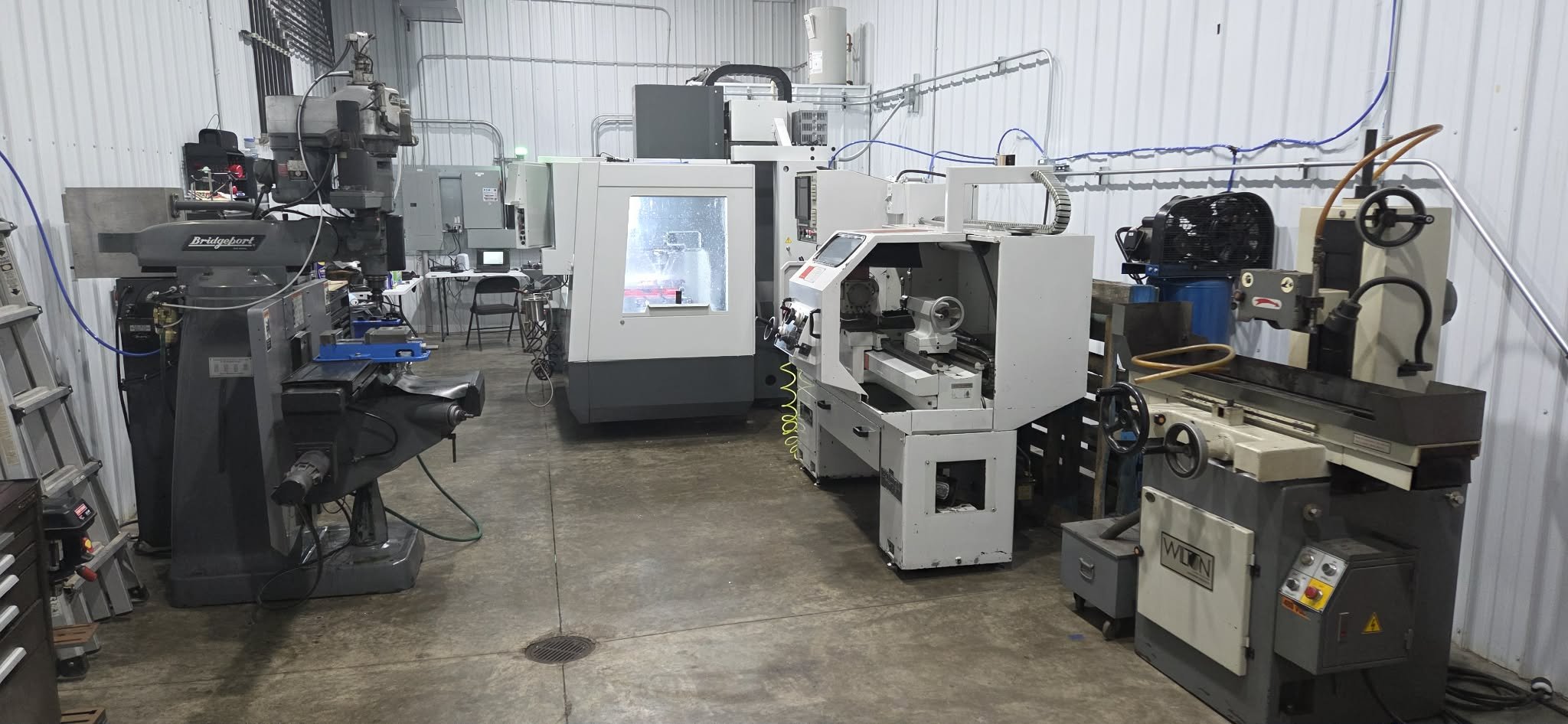 Industrial workshop with various CNC machines and equipment on concrete floor, metal walls, and a ladder on the left side.