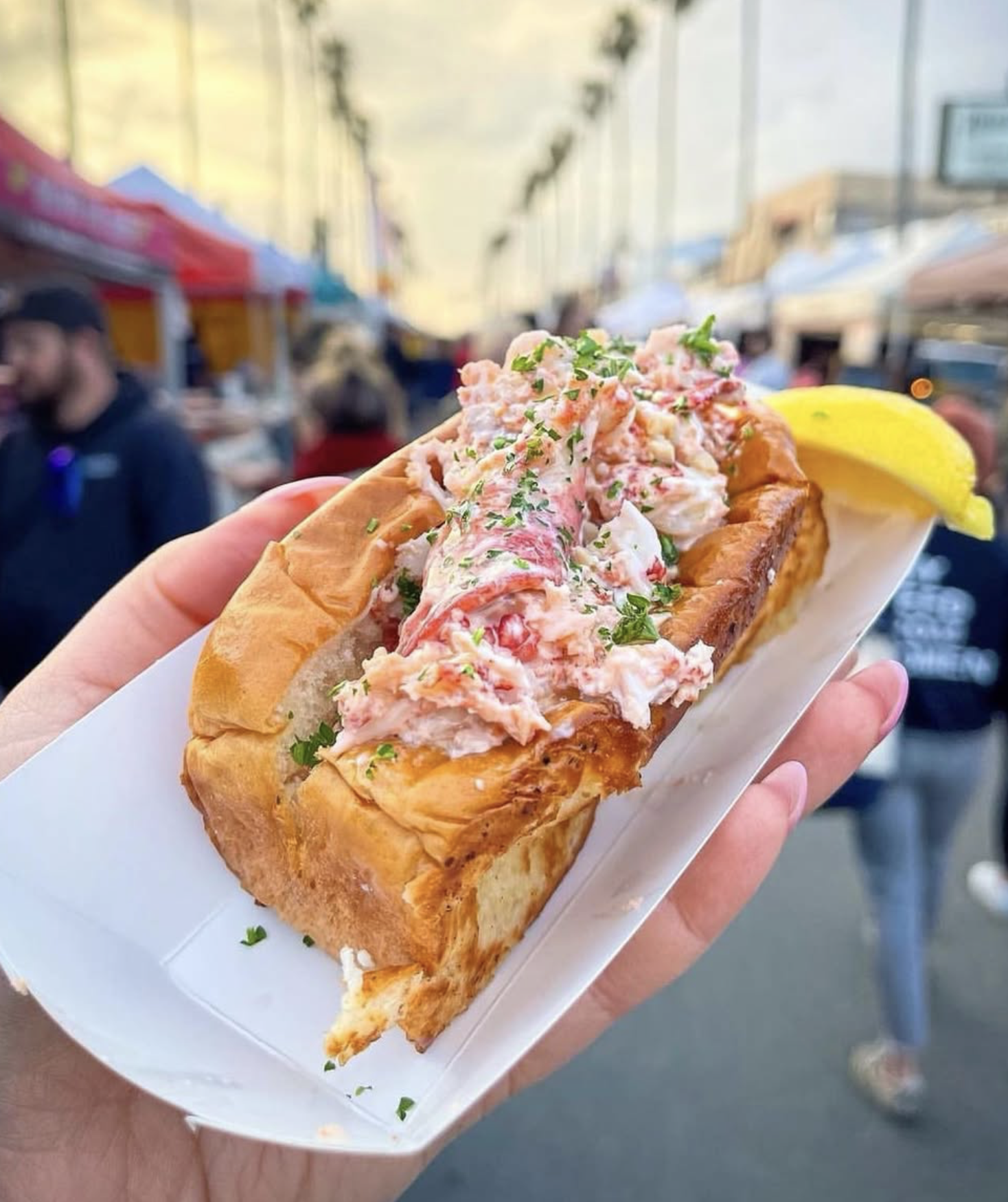 A hand holding a lobster roll with lobster meat, herbs, and mayonnaise in a toasted bun, with a lemon wedge on the side, at a food market with blurred people and tents in the background.