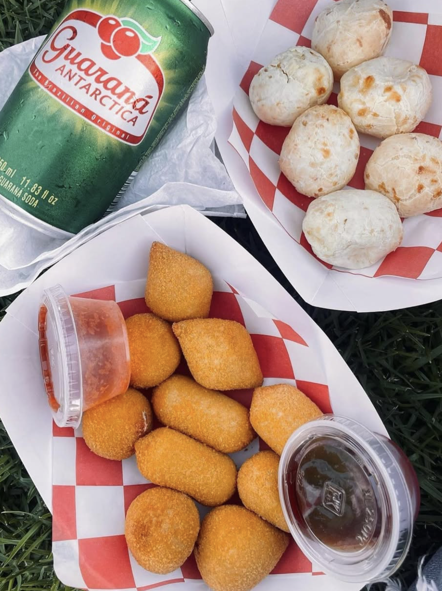 A picnic spread with a can of guaraná soda, a basket of fried cheese balls, a basket of soft bread rolls, and two small cups of dipping sauces, all arranged on checkered paper on grass.
