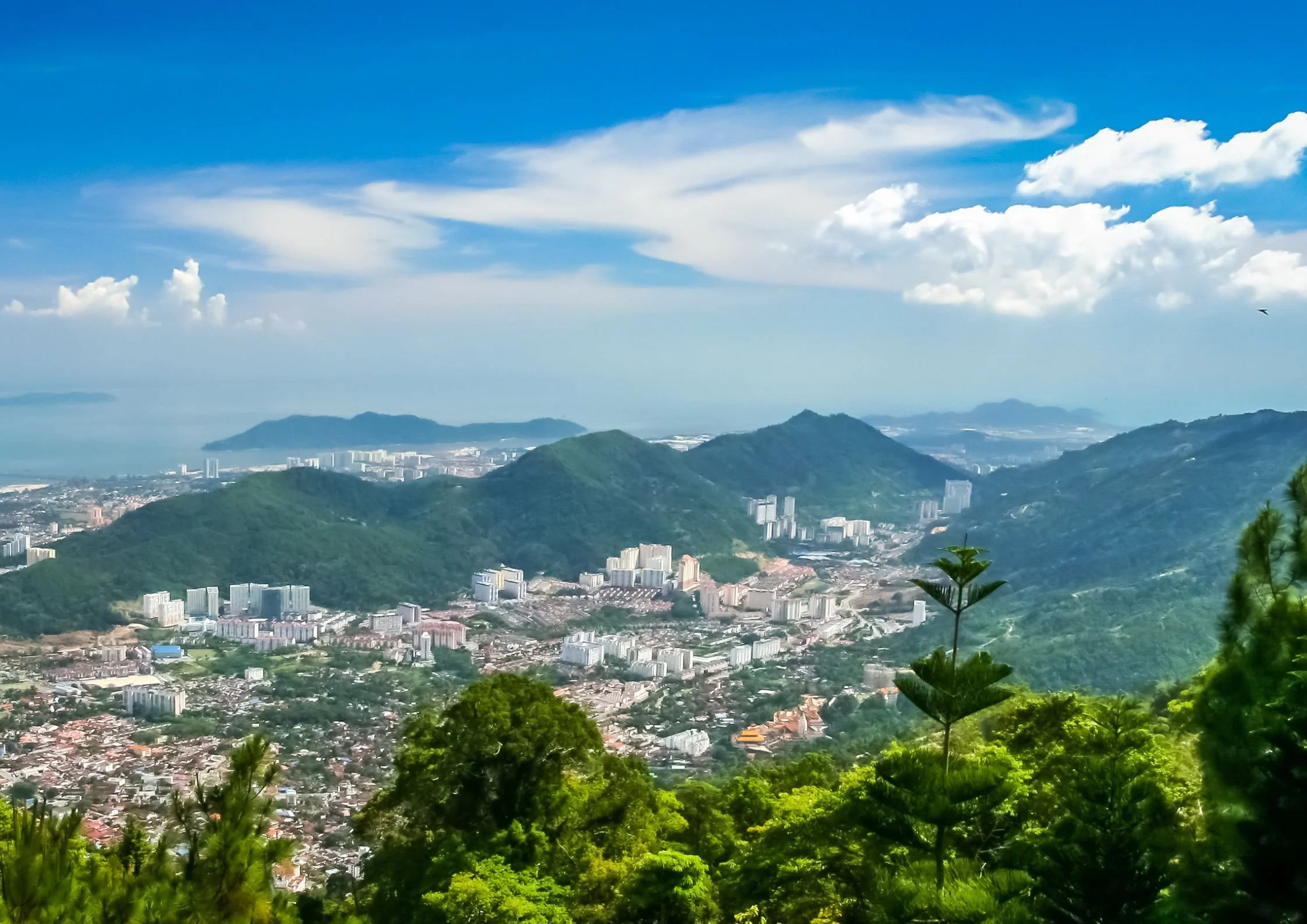 A panoramic view of a city nestled in green mountains with high-rise buildings, a coastline in the background, and a partly cloudy blue sky above.