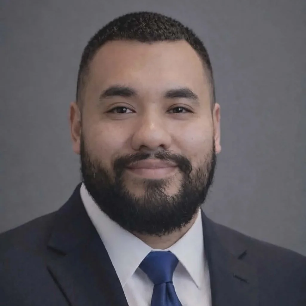 Professional headshot of a man wearing a dark suit, white shirt, and blue tie, with short hair and a beard, against a gray background.