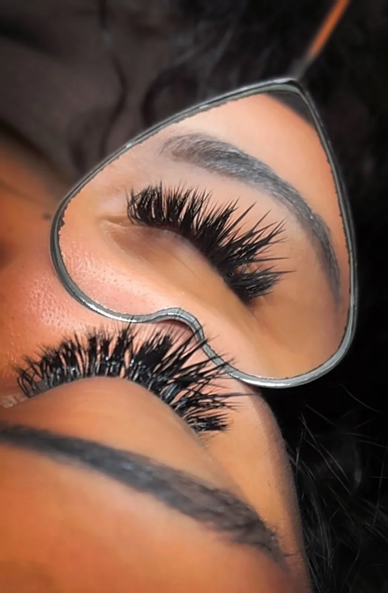A person receiving eyelash extension treatment with a technician applying false eyelashes using tweezers, wearing eye pads under their eyes.