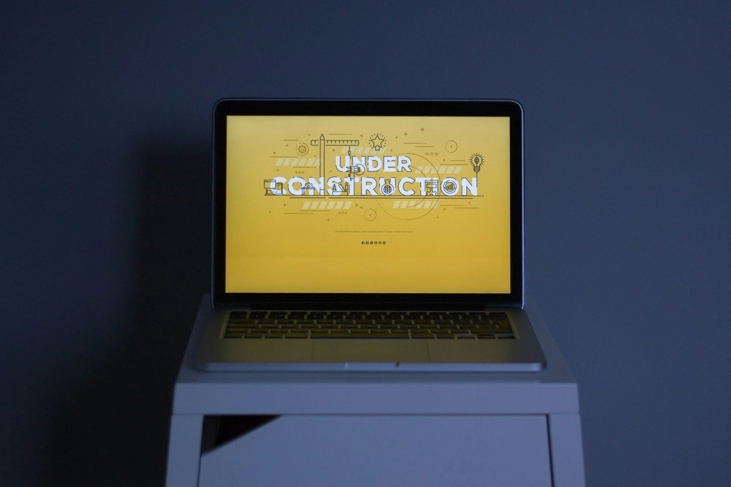 A laptop displays a yellow screen with the words 'Under Construction' in large text, surrounded by construction-themed icons, such as a ruler, pencil, lightbulb, and gears, indicating a website or project that is currently being built or updated.