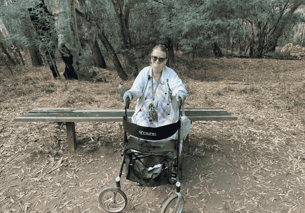 Woman with sunglasses sitting in a wheelchair on a trail in a wooded area, with a bench behind her.