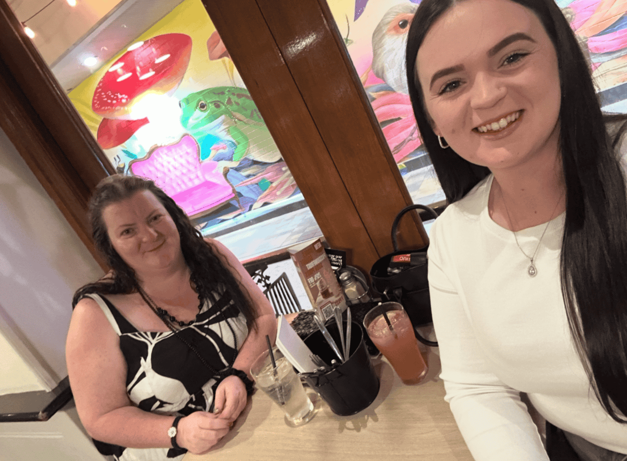 Two women sitting at a table in a restaurant with colorful mural art of animals including frog, bird, and mushrooms in the background. One woman with long dark hair, wearing a white top, is smiling at the camera. The other woman with curly dark hair,