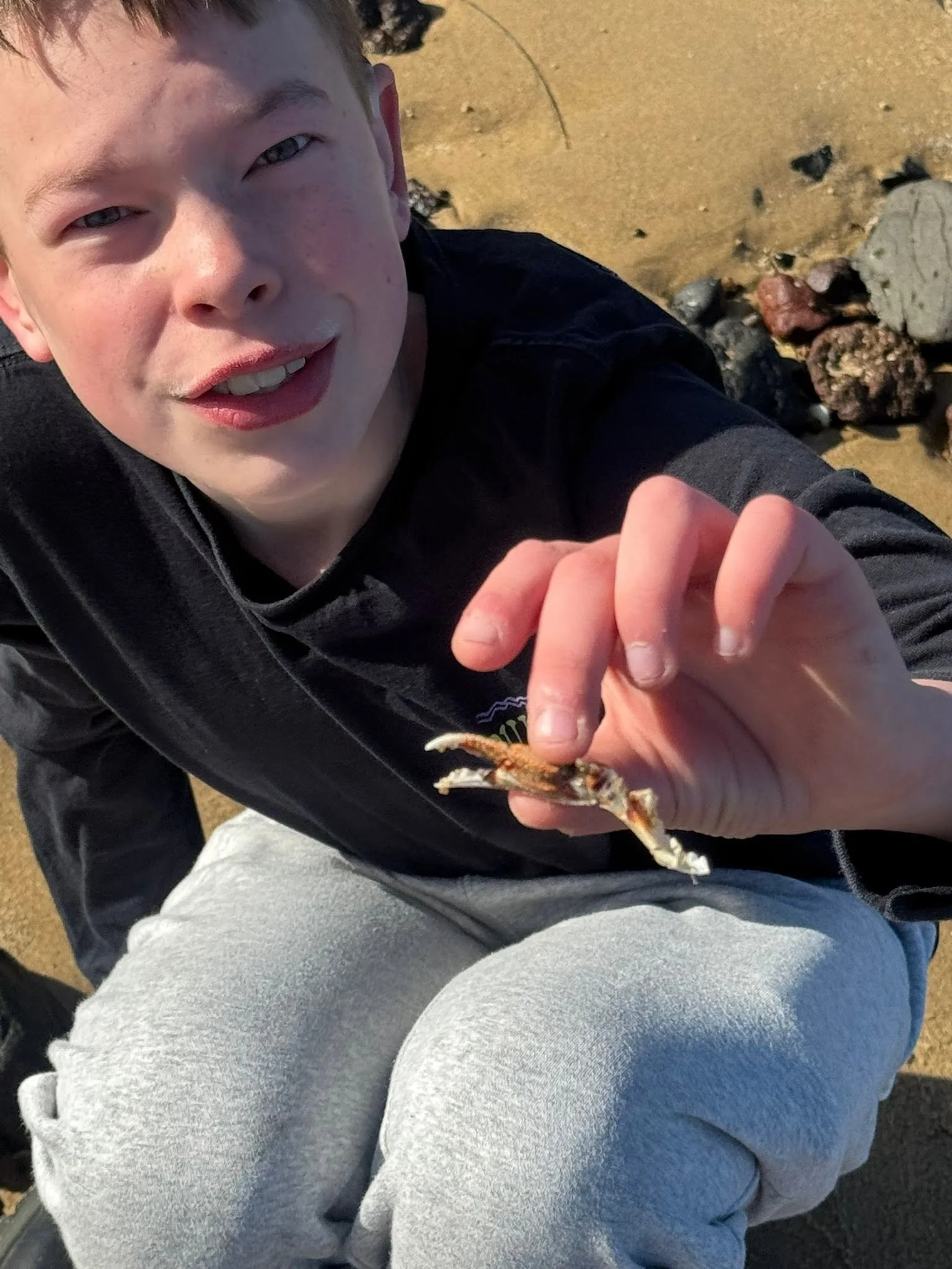 A close-up of a smiling person holding a small fish by the tail, while sitting on a sandy beach with rocks in the background.
