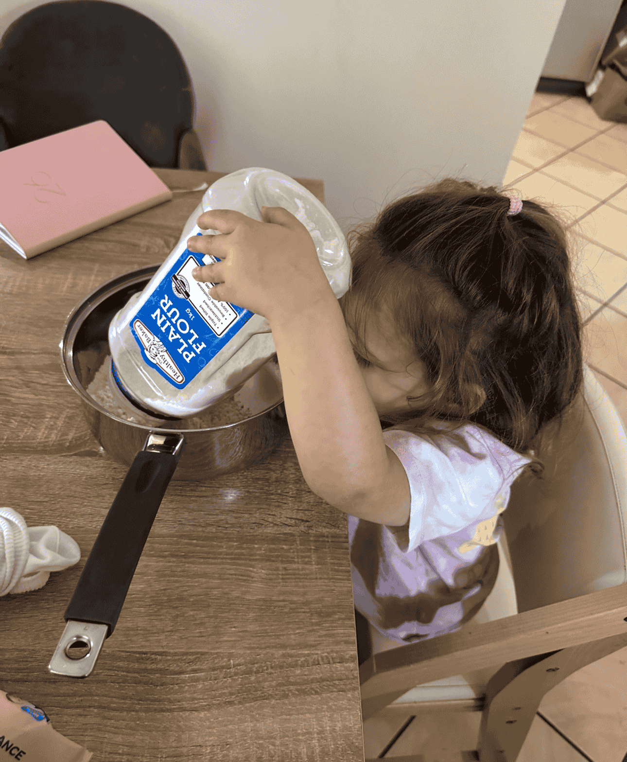 A young girl with brown hair in a ponytail is pouring heavy whipping cream from a large package into a metal saucepan on a wooden table.