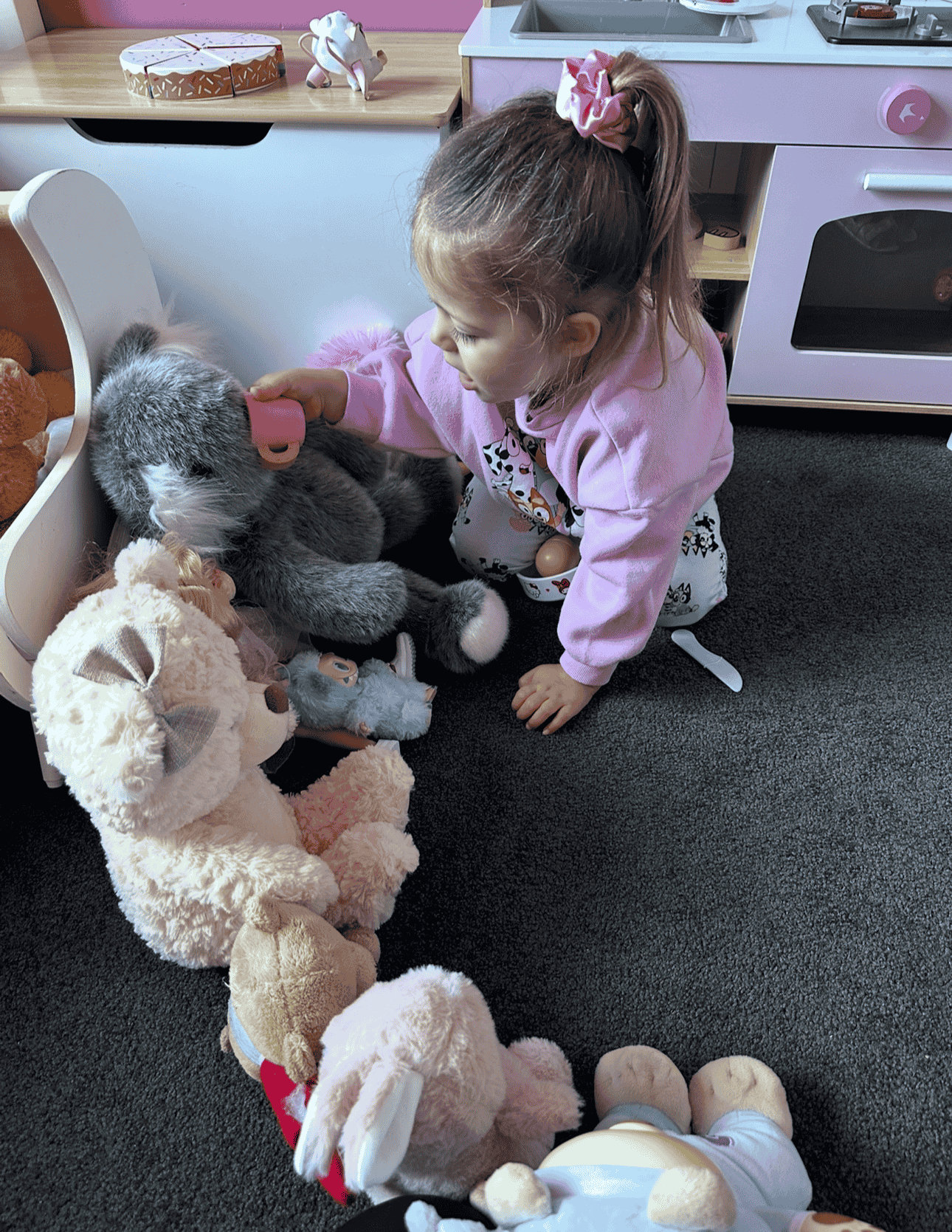 A young girl with a pink hair scrunchie and pajamas is playfully pretending to groom a large stuffed koala bear toy with a pink hairbrush while surrounded by other plush toys on a black carpet in a play area with toy kitchen appliances in the background.
