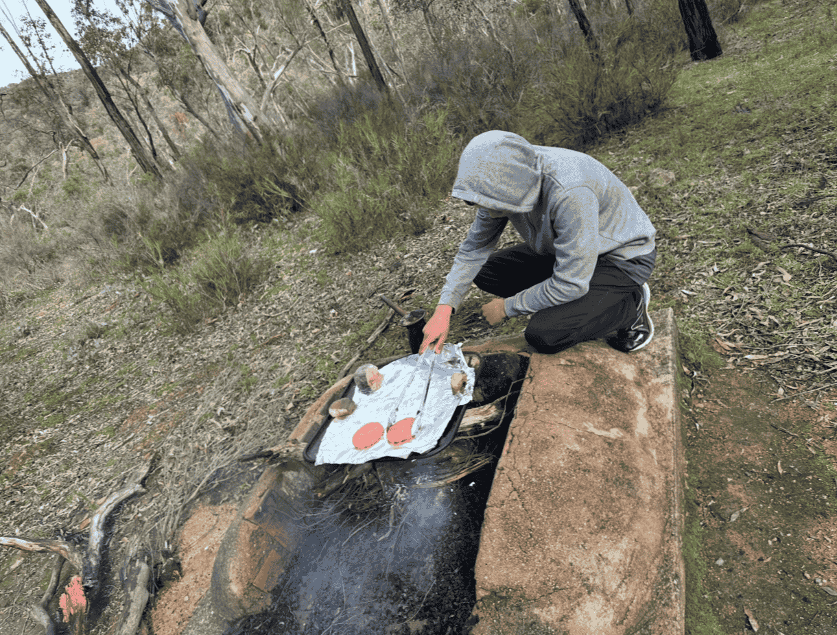 A person wearing a gray hoodie and black pants is crouching on a brick ledge next to a small campfire. The person is lifting a piece of aluminum foil covering three baked potatoes on a tray, which is placed on top of the fire. The setting is outdoors