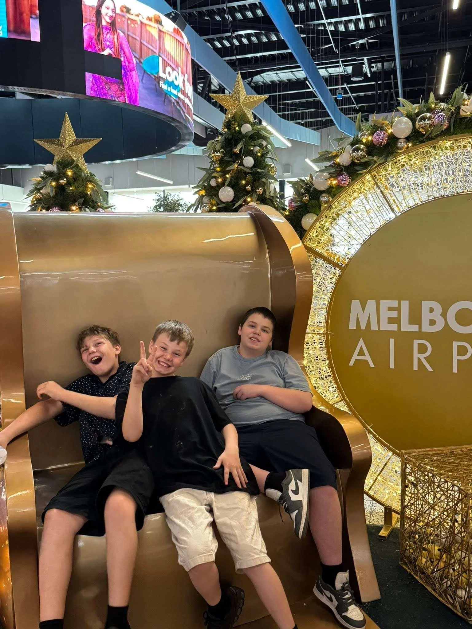 Three boys sitting on a gold-colored bench with Christmas trees decorated with ornaments and lights behind them at Melbourne Airport.